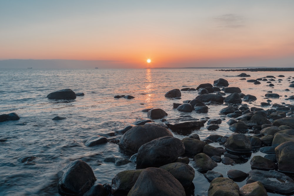 the sun is setting over the ocean with rocks in the foreground