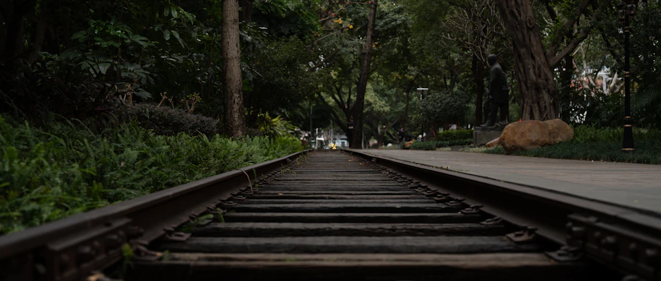 The image shows a long, straight train track winding through a lush park setting. The track is made of wood and runs from the foreground to the background, disappearing into the distance. On either side of the track are trees with green leaves, providing a natural backdrop for the scene. In the middle ground, there's a statue situated on a small hill, adding an artistic element to the landscape.