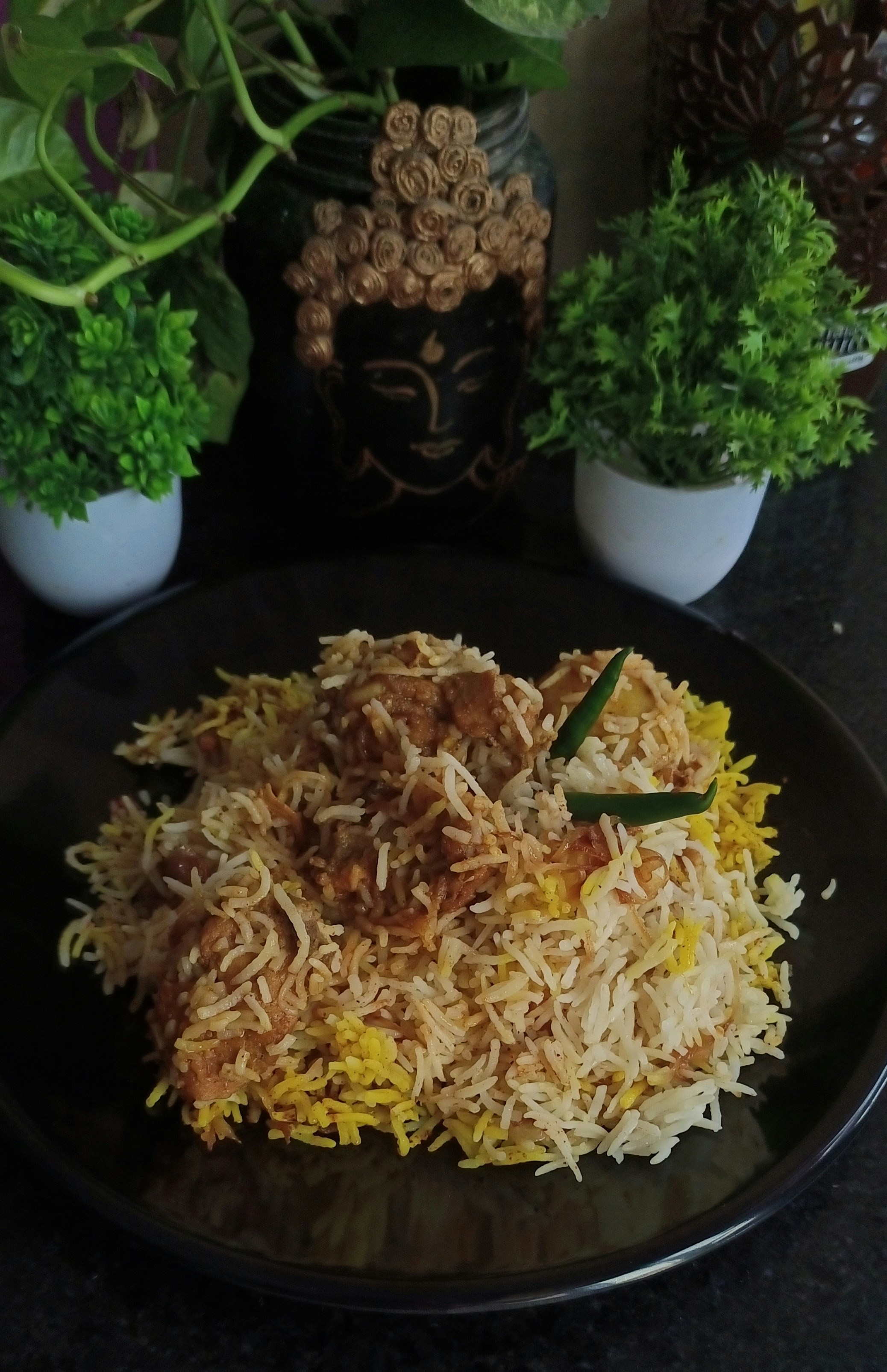 A black plate of biryani, white and saffron-yellow rice with pieces of meat and green chillies