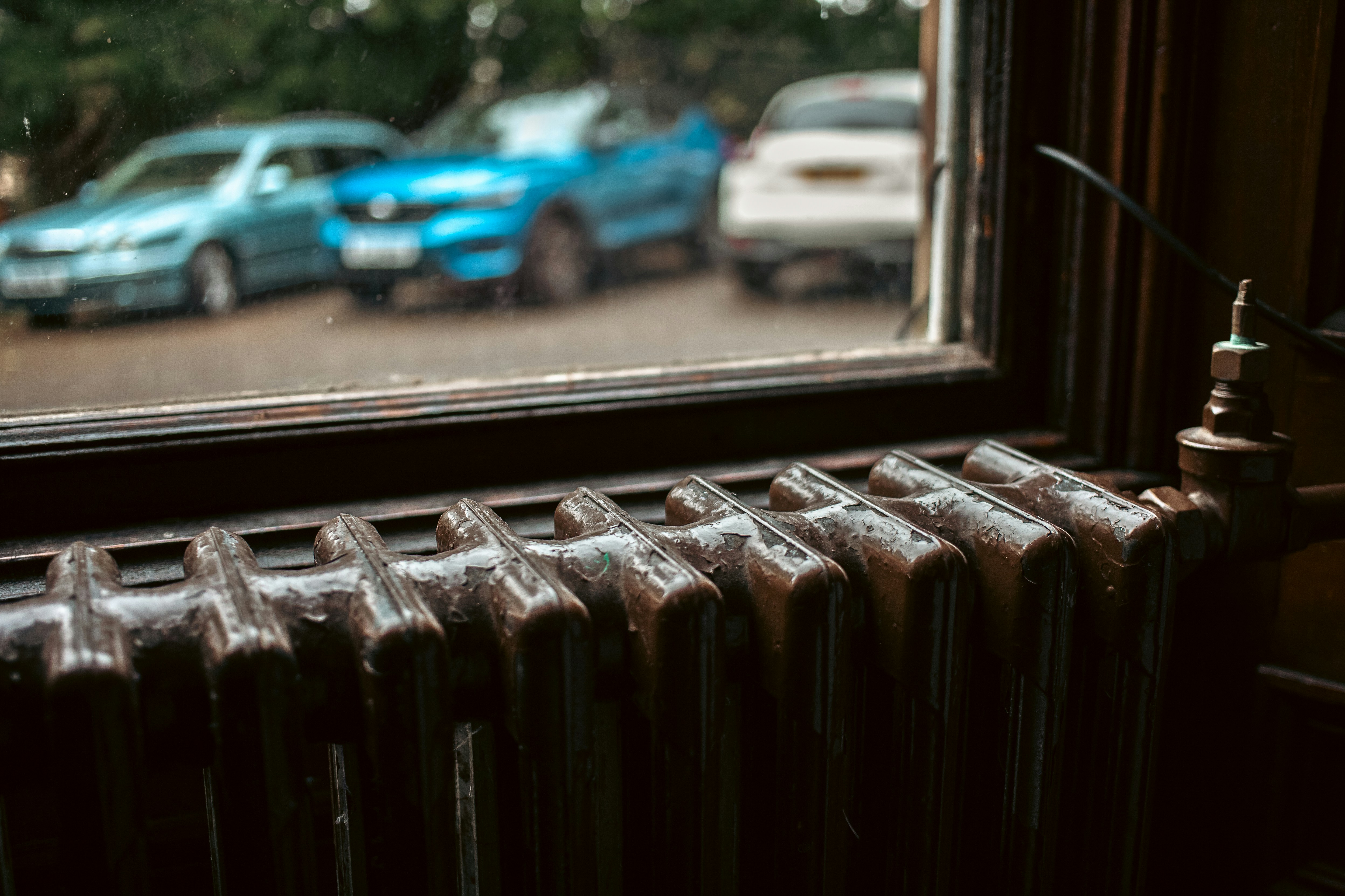 Un radiateur en fonte sous une fenêtre donnant sur un parking, à l'approche de la saison de chauffe