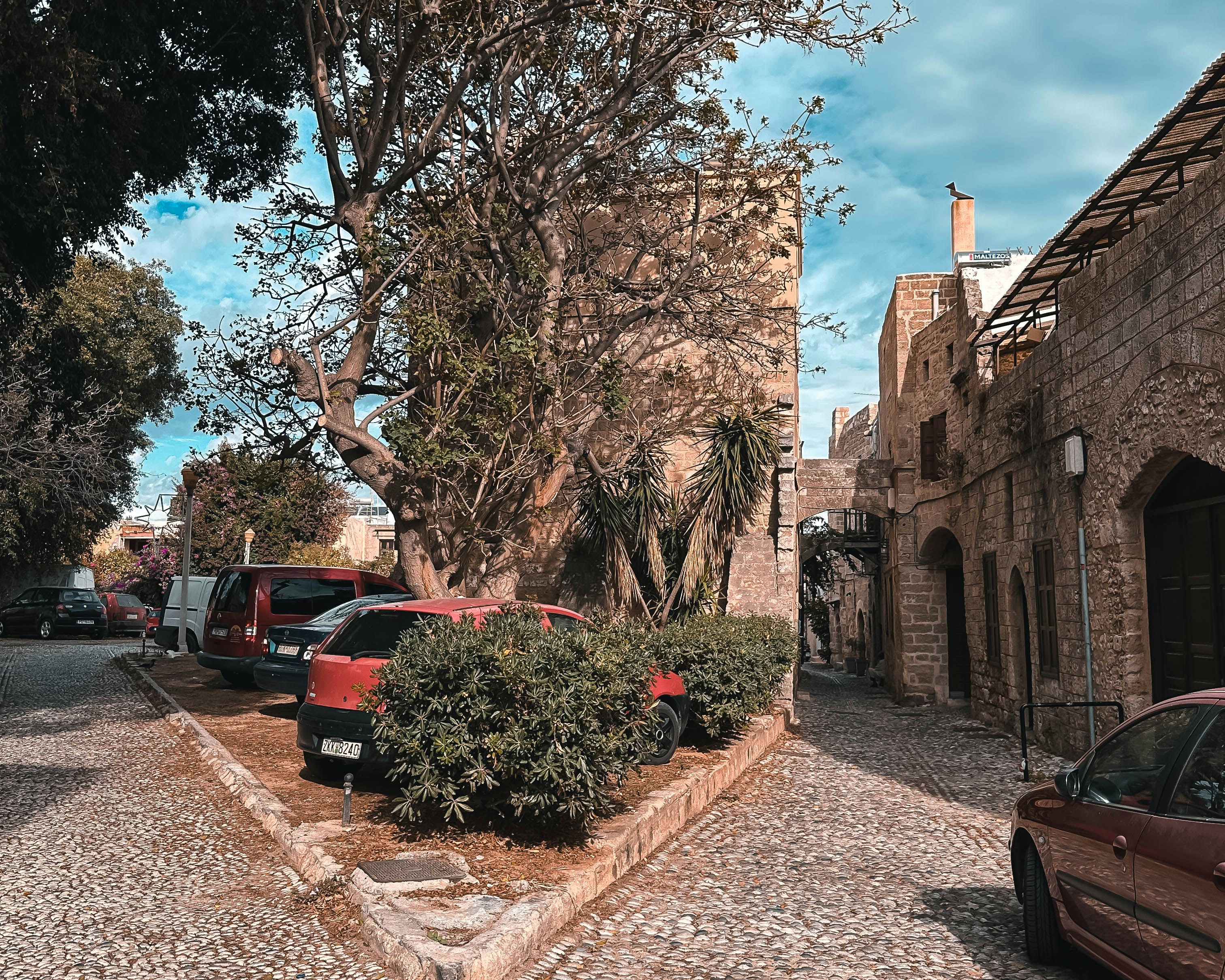 a car parked on the side of a road next to a tree, 📸 Lose yourself in the maze of cobbled streets and hidden alleys that wind through the historic heart of Rhodes. Each corner reveals a new treasure – from quaint cafes to artisan shops, and colorful bougainvillea cascading down ancient stone walls.