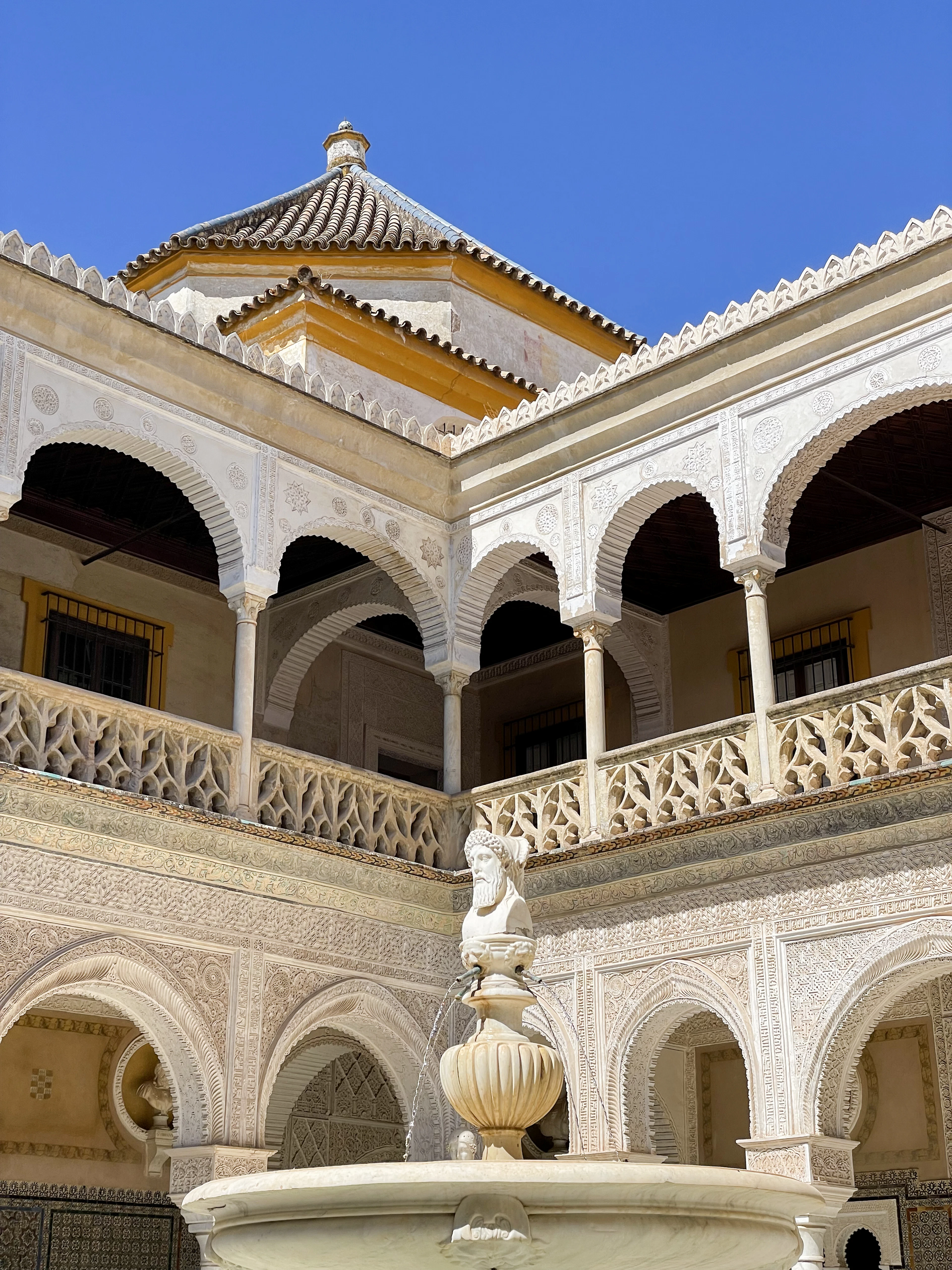 The Mudejar patio of the Casa de Pilatos in Seville in full sun: a white plasterwork arcade on slender marble columns above a lacework balustrade, with the marble fountain and its bearded bust in the foreground.