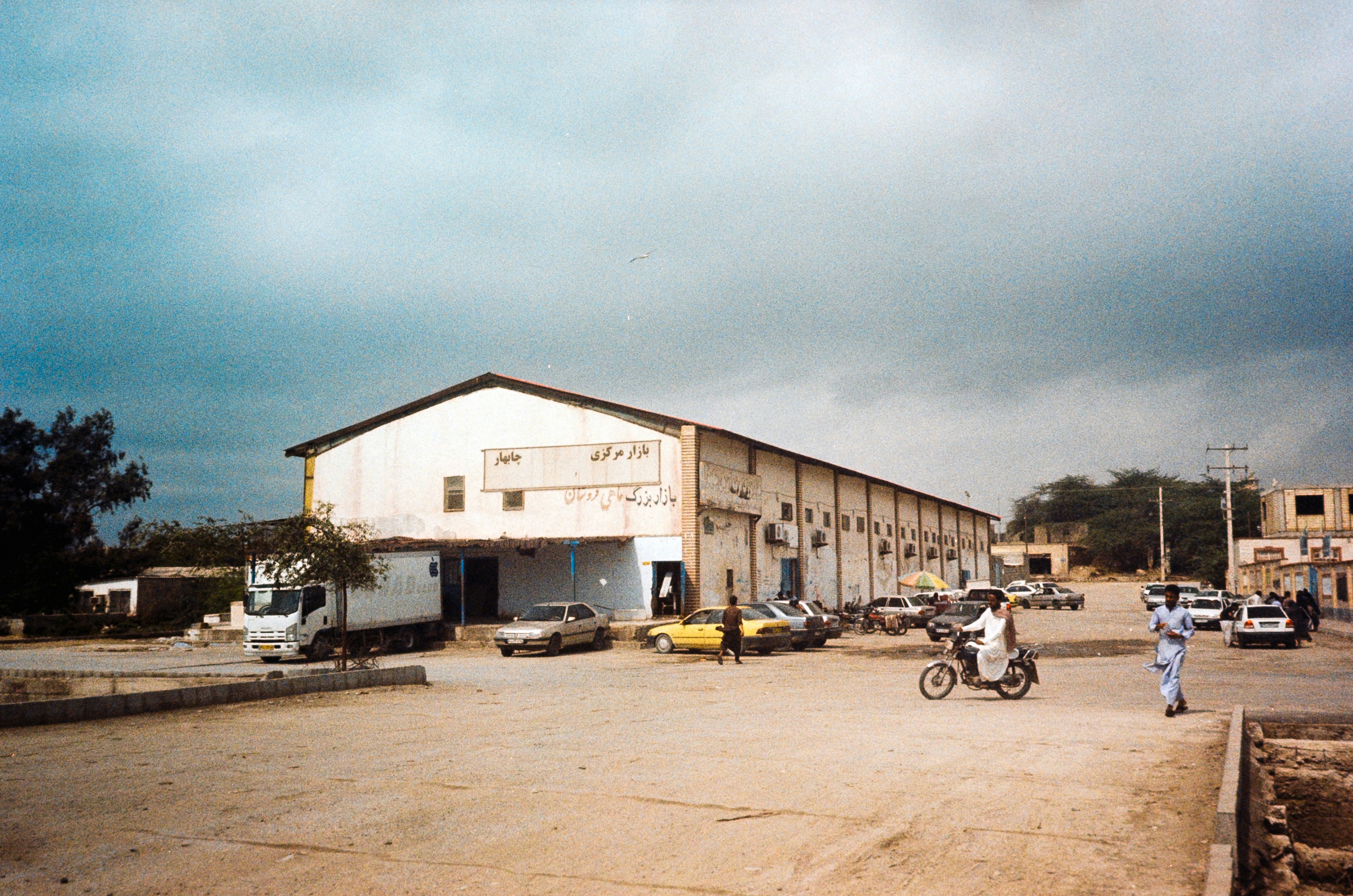 (The people in this photo have the consent to be in it) A bustling seafood market, housed in a large building, surrounded by parked cars and motorcycles, with people milling about outside.