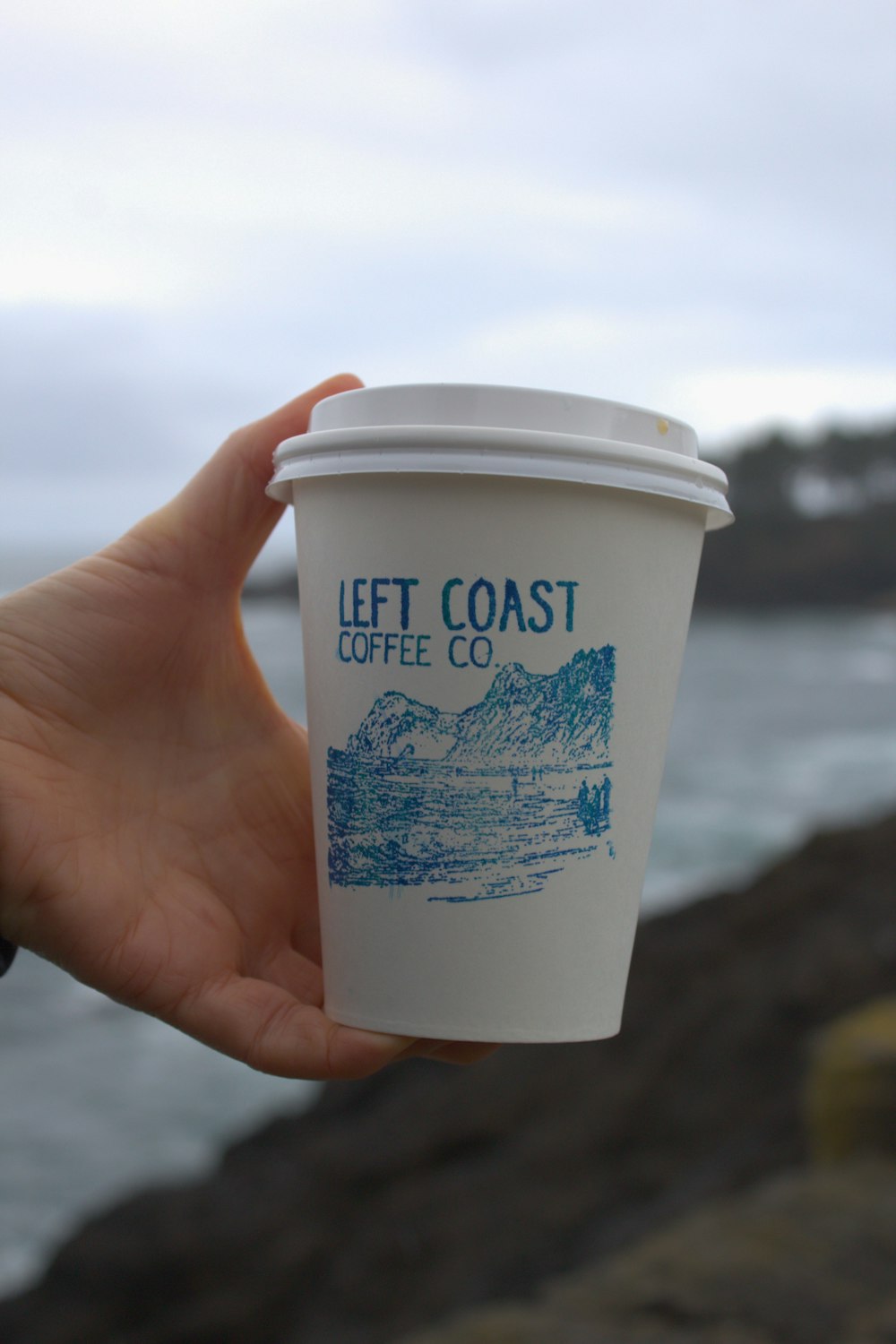a hand holding a cup of coffee near the ocean