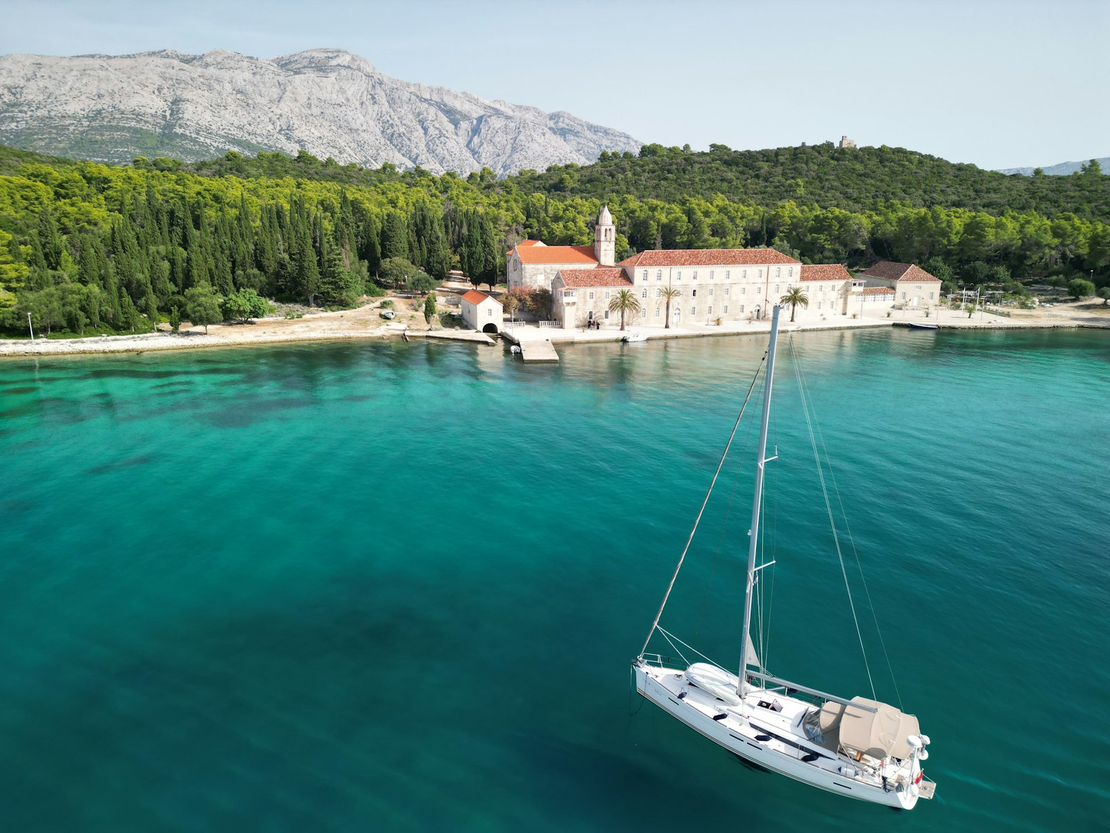 A sailboat anchored in turquoise water off a cypress-lined island monastery near Korčula, with mountains behind