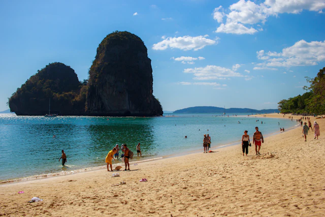 A tall limestone karst tower rises straight out of the turquoise water at the edge of a sandy beach where people wade and walk.