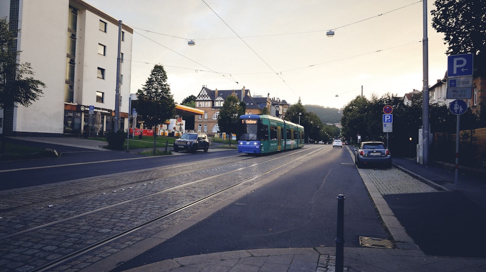 a blue and green train traveling down a street next to tall buildings