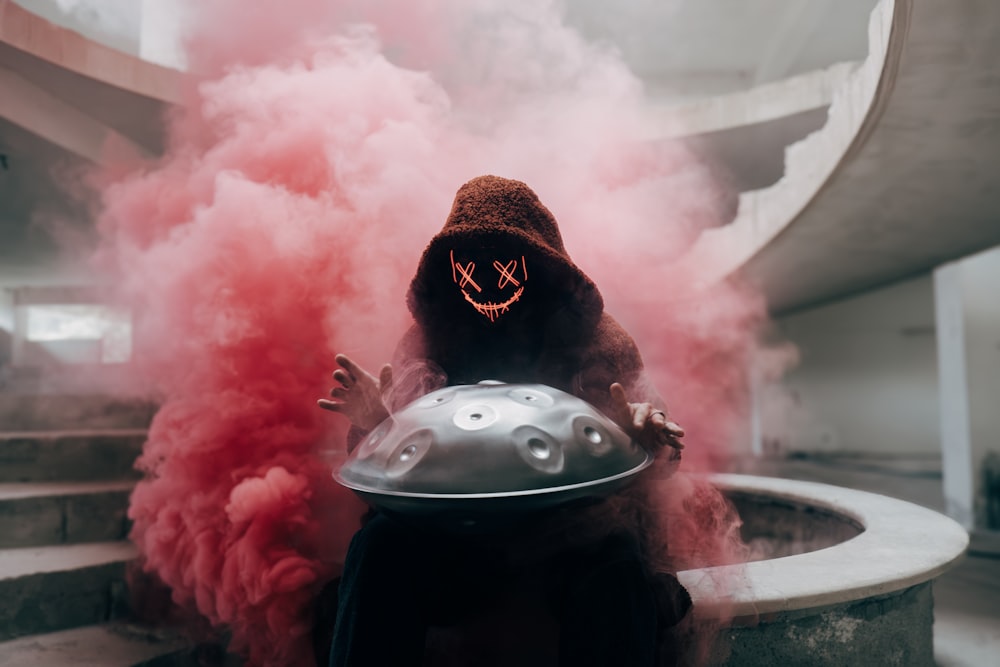 a person wearing a hood and holding a helmet in front of a cloud of smoke