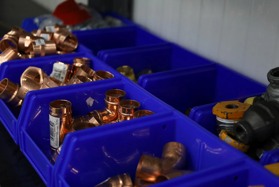 The image shows a collection of copper pipes and fittings neatly organized within blue plastic bins on a white shelf against a gray wall backdrop. The bins are arranged in two rows, with three bins on each side of the shelf. Each bin contains various sizes and types of copper pipes and fittings, including larger pipes at the bottom row and smaller ones towards the top.