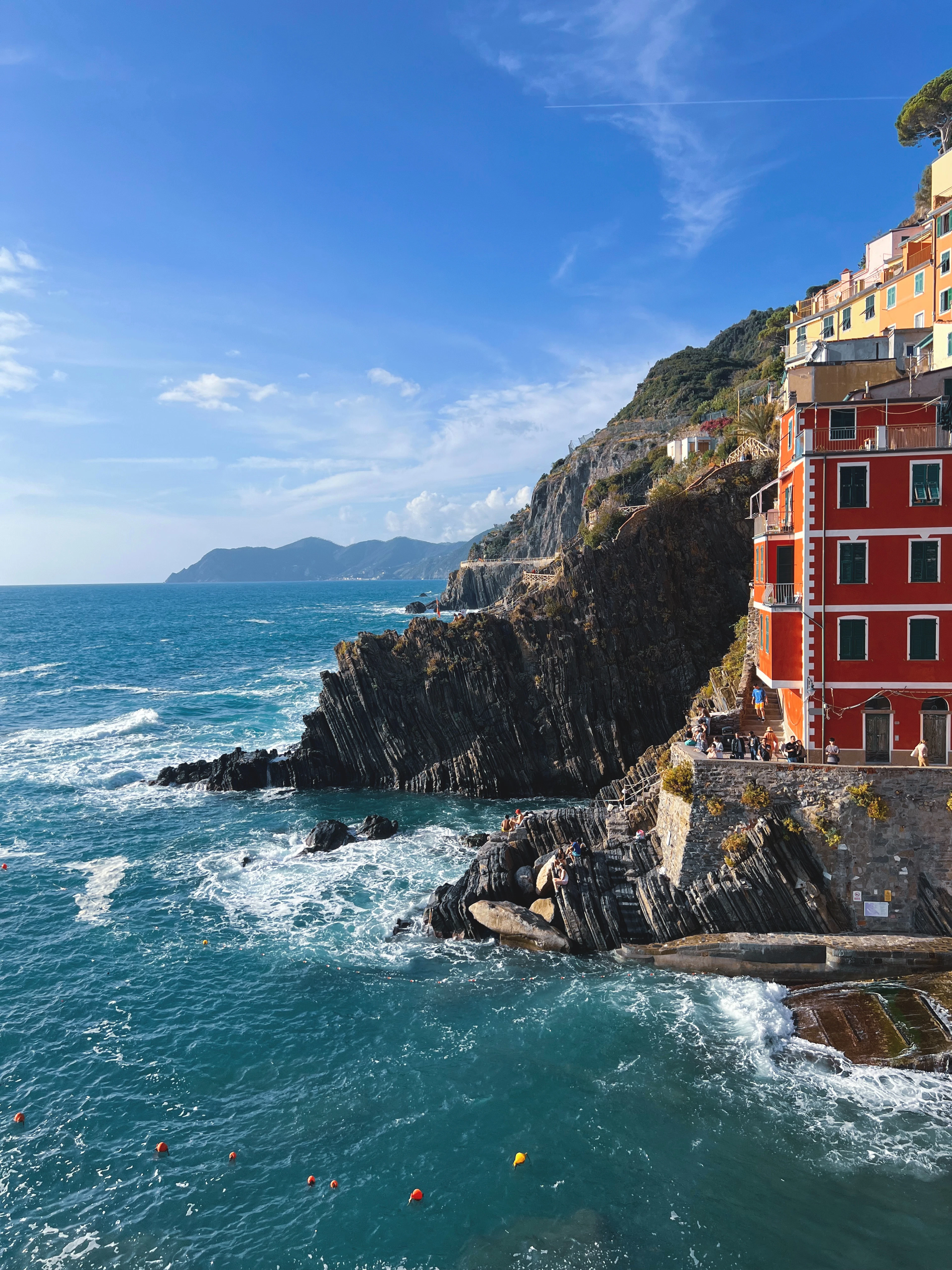 A tall red house at the edge of Riomaggiore standing above dark layered cliffs, with surf breaking white against the rock and the turquoise Ligurian Sea stretching to distant headlands under a blue sky