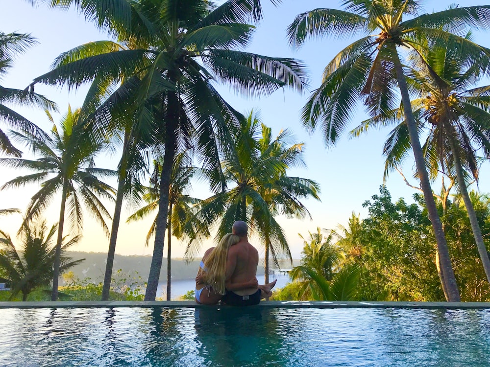 a man sitting on the edge of a swimming pool surrounded by palm trees