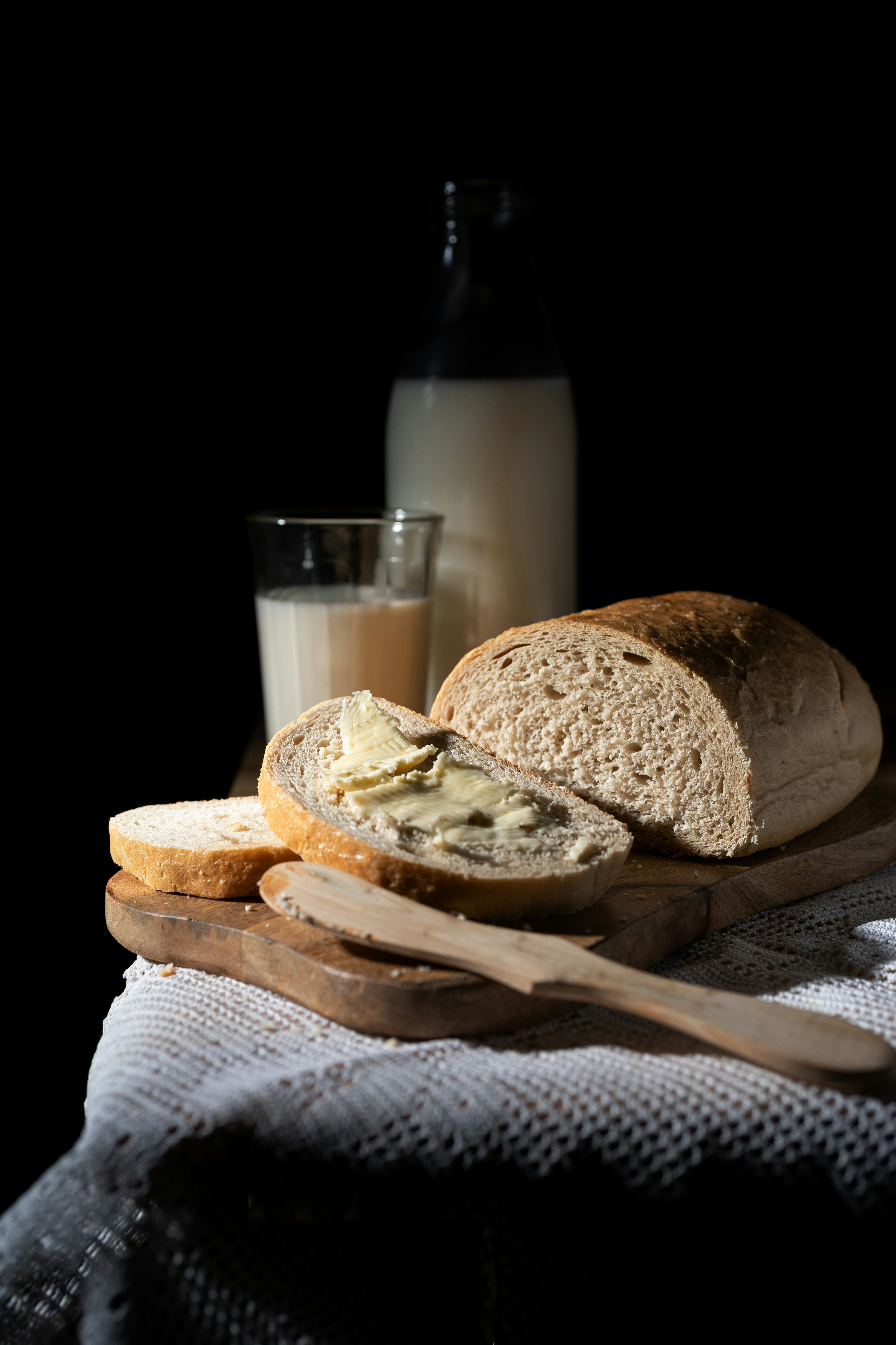 Pieza de pan cortada en rodajas sobre una tabla de madera, un vaso con leche y una botella detrás con leche.
