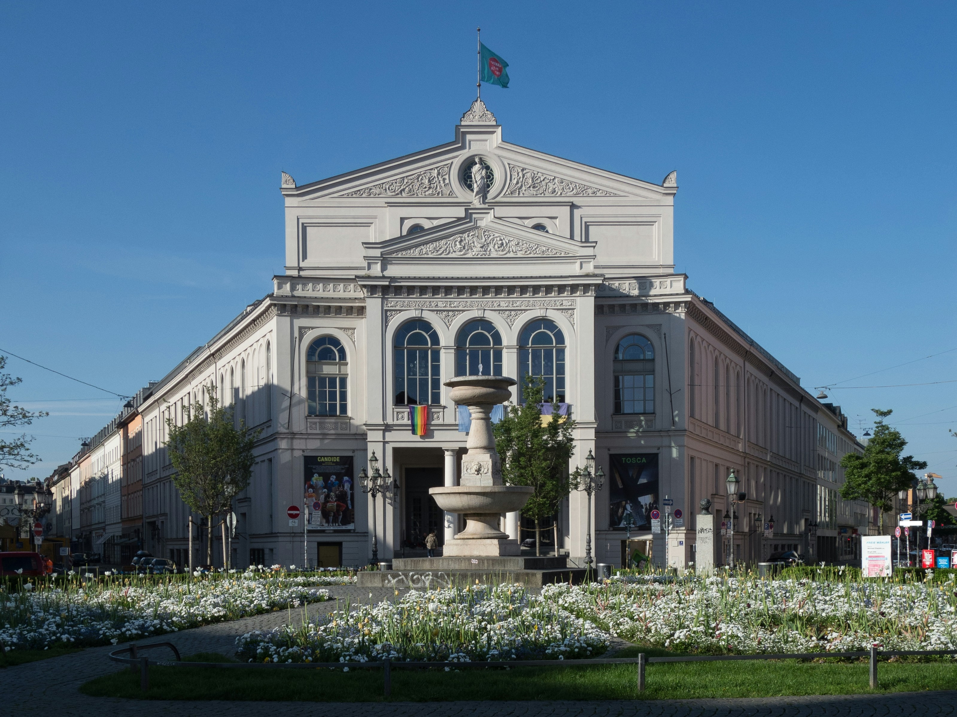 A grand Munich opera-theatre building with a fountain in its forecourt under a blue sky