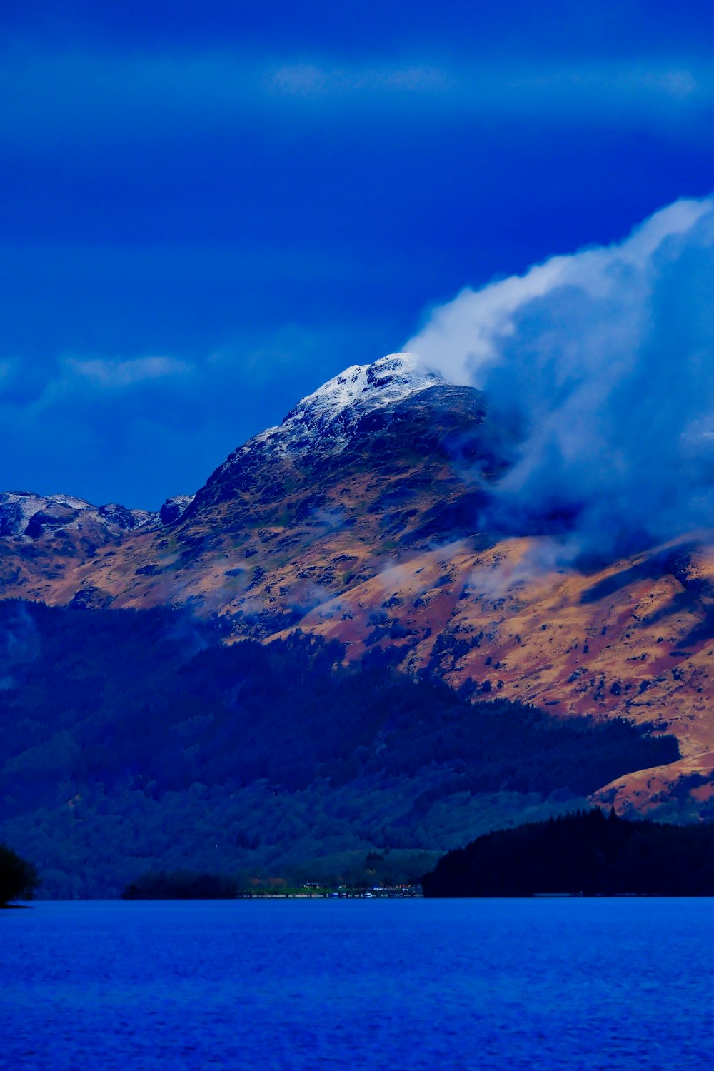 a mountain covered in snow and clouds next to a body of water