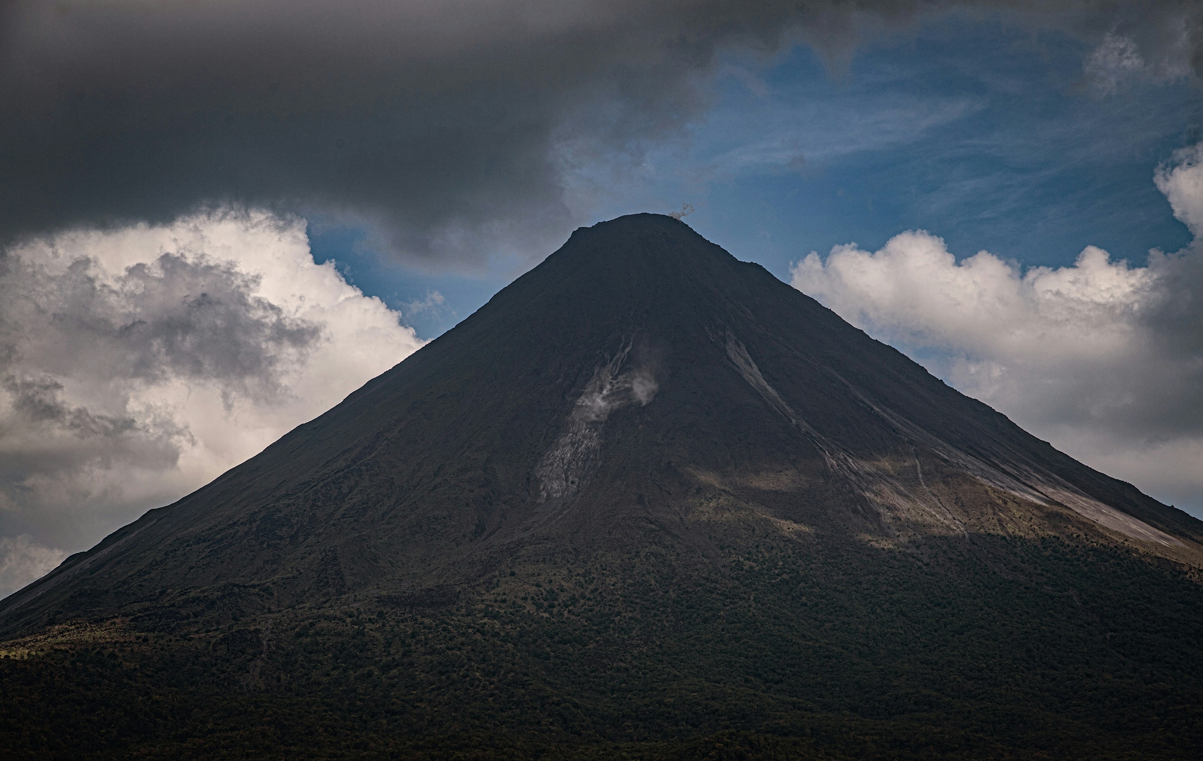 a very tall mountain under a cloudy sky, This image captures the Arenal Volcano in Costa Rica with its conical shape and a visible trail of smoke at its summit. The volcano stands against a dramatic sky, with clouds gathering around the peak, hinting at the geothermal activity within. Its forested slopes and the rugged terrain around it are also visible.