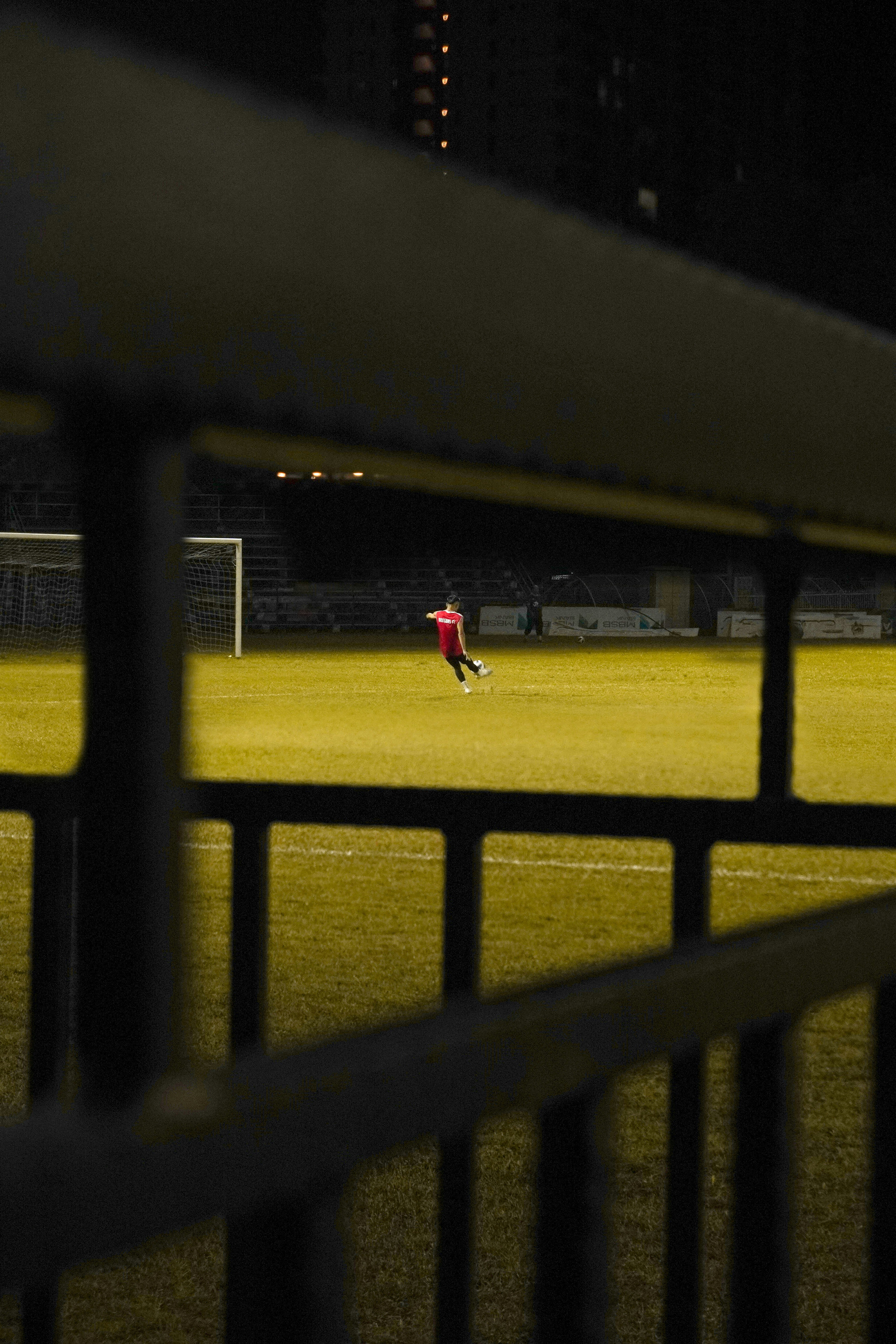 Looking through a railing at an empty soccer stadium. A light-skinned player in a red jersey kicks a ball toward the goal