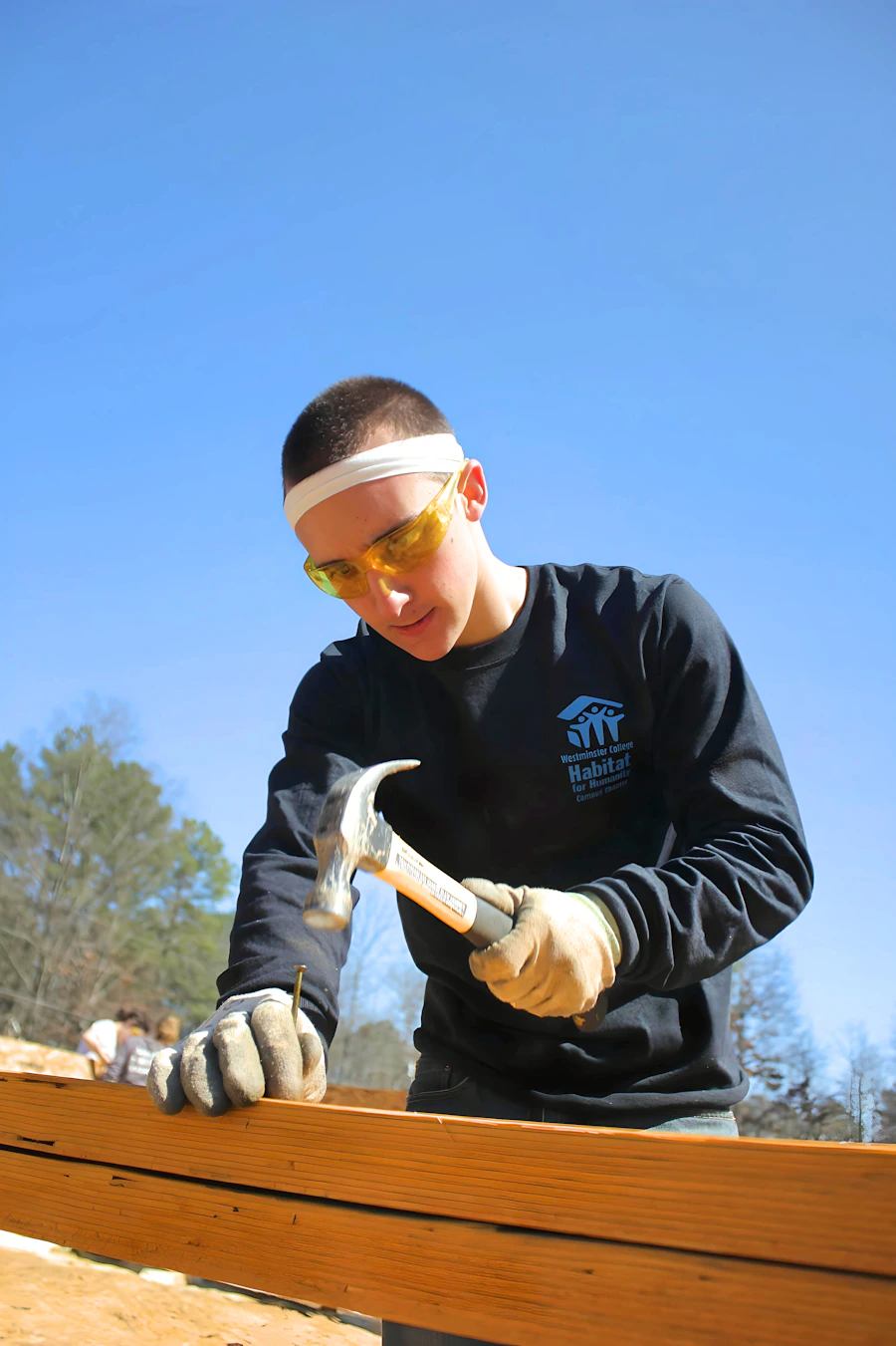 A young man in safety glasses and gloves driving a nail with a hammer