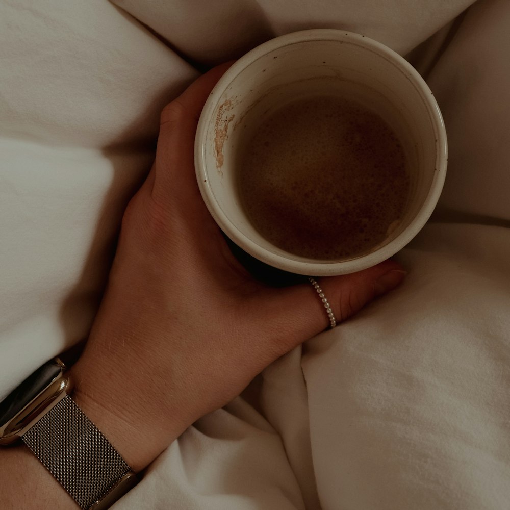 a person's hand holding a cup of coffee on a bed
