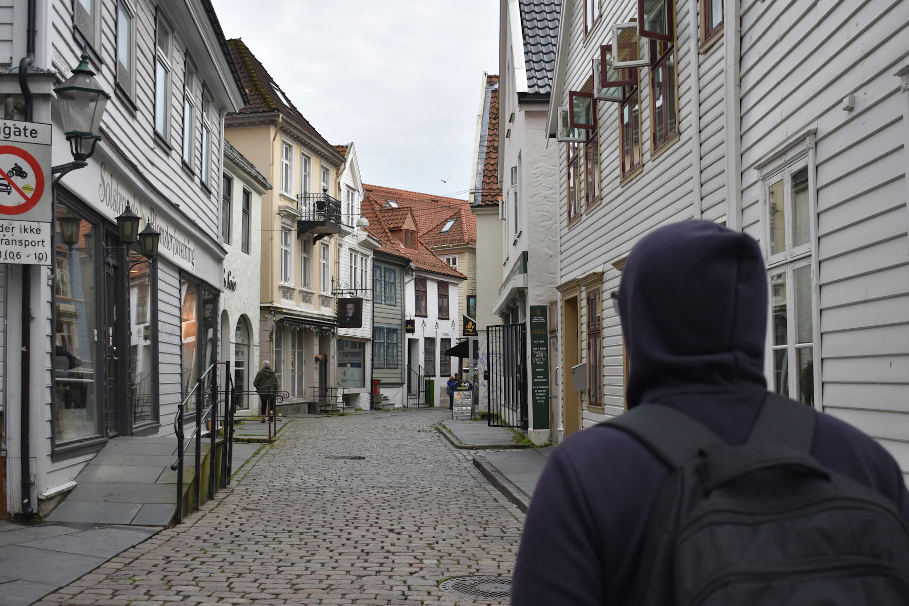 Seen from behind, a traveler in a hooded jacket with a backpack on a cobbled street of white wooden houses
