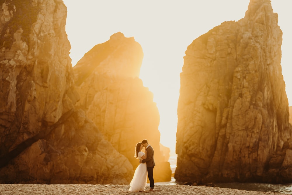 a bride and groom standing on a beach in front of large rocks