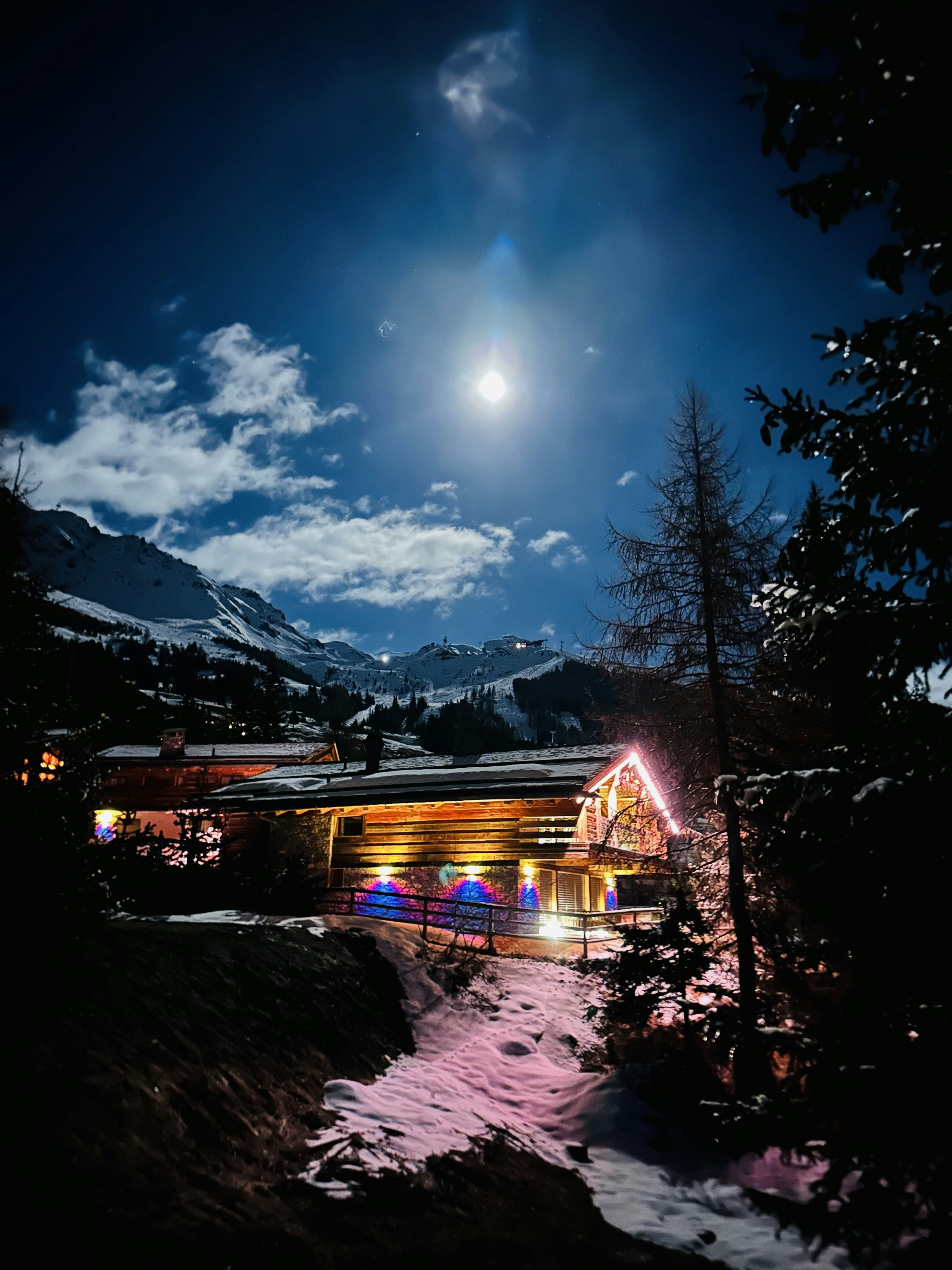 Contemporary mountain lodge at night, warm interior light glowing through large windows, snow-dusted pine trees, dark moody sky