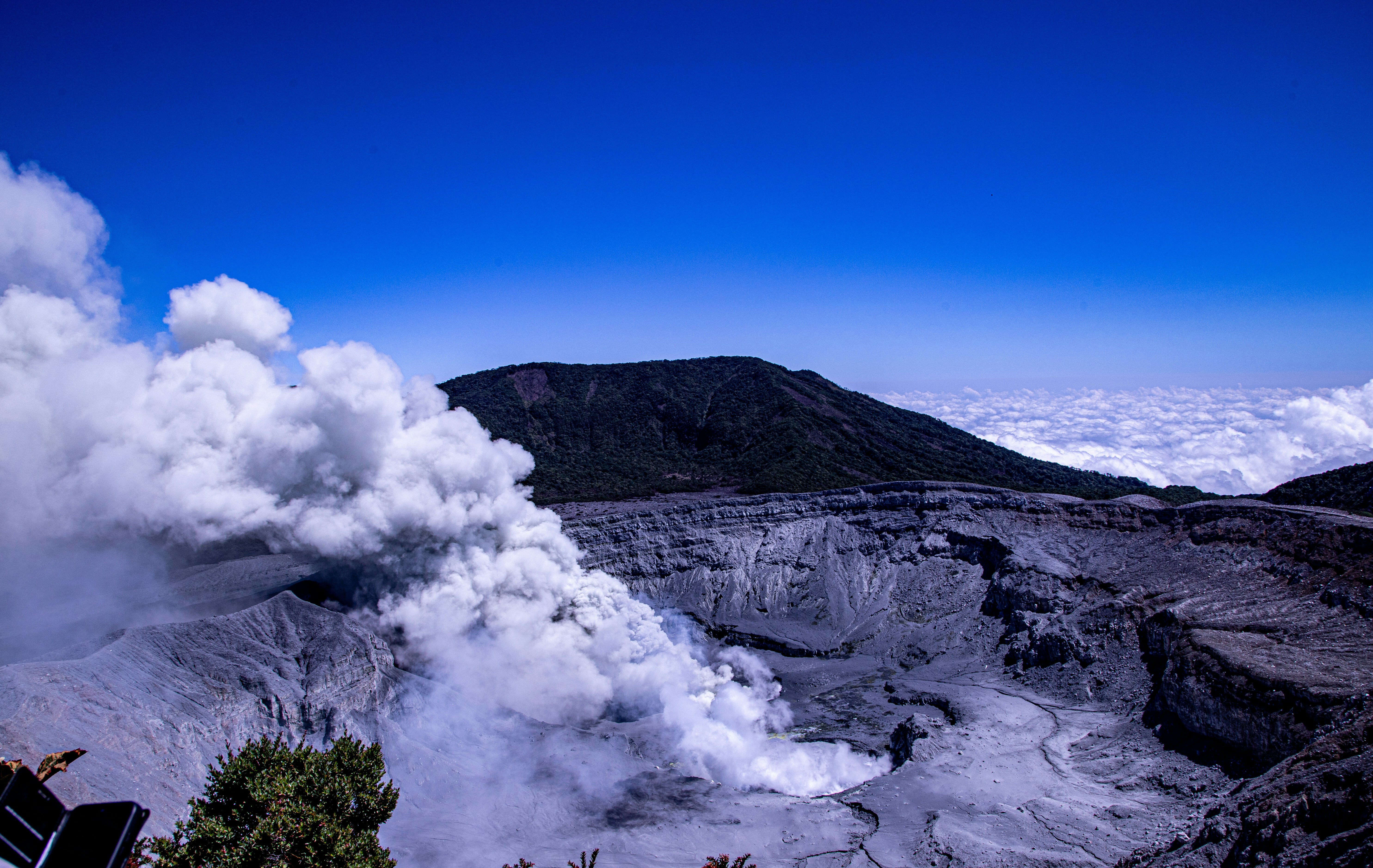 a large plume of smoke coming out of a mountain, A panoramic view of the Poás Volcano crater, with plumes of white steam rising from its geothermal vents, set against a backdrop of a cloud-filled sky and surrounding rugged terrain.