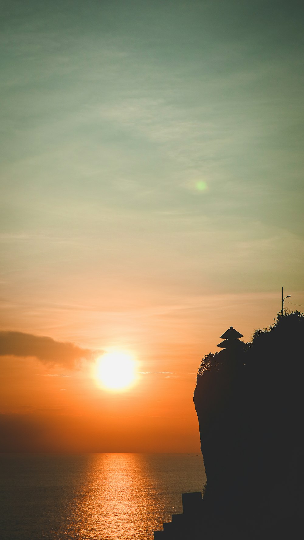 the sun is setting over the ocean with a small hut on the cliff