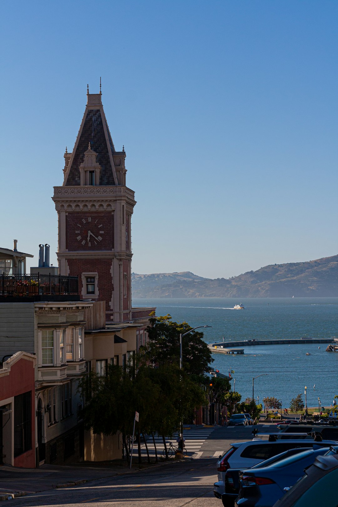 This image offers a picturesque view of the San Francisco skyline, crowned by the ornate clock tower against the blue expanse of sky and sea. The steep incline of the city street, lined with classic architecture, invites viewers to ponder the balance of urban development with natural beauty a theme ripe for exploration on lifestyle or travel blogs. The clock tower, with its timeless design, serves as a focal point that anchors the city’s historical significance within its modern context.