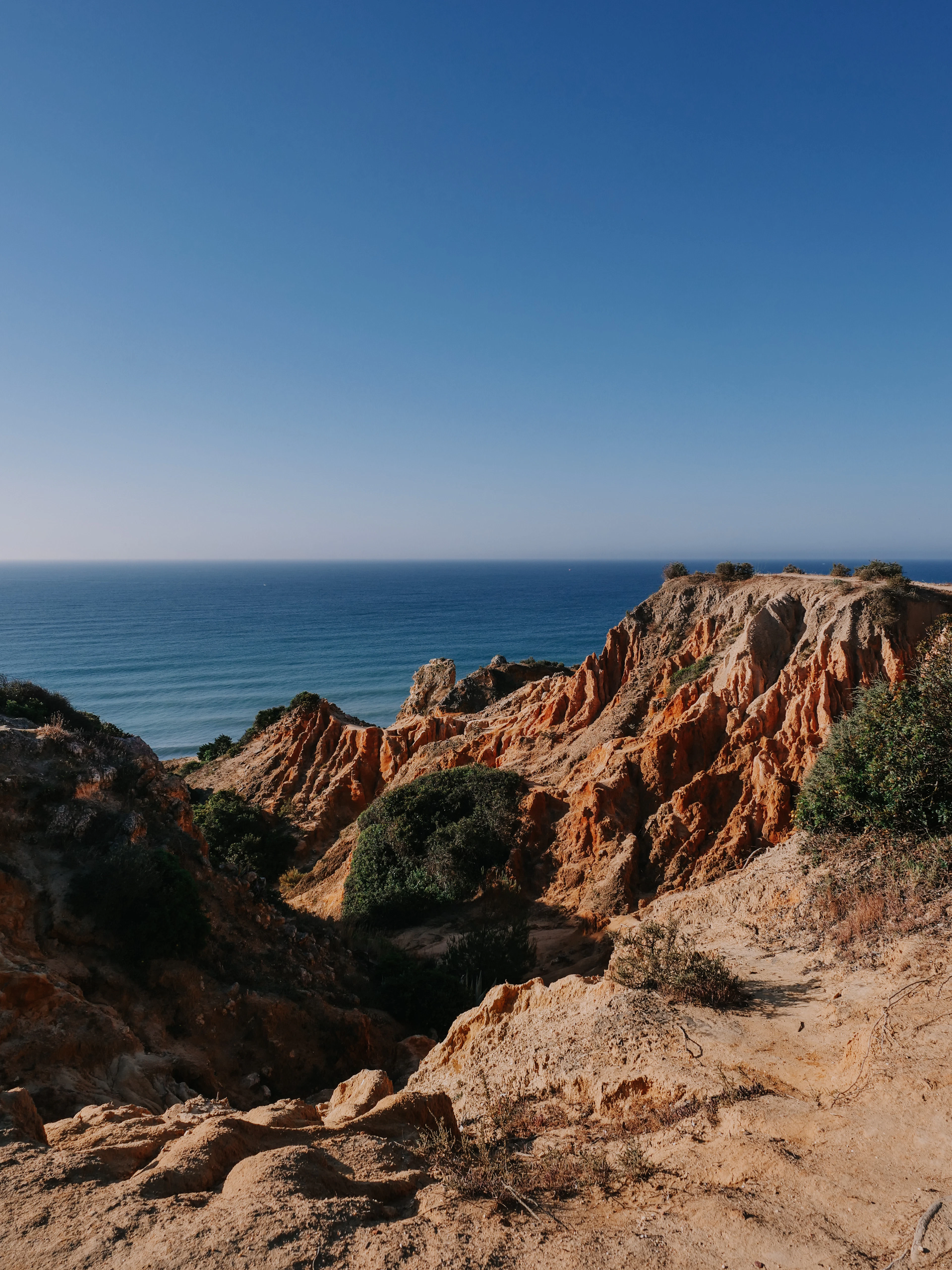 Sunlit red and orange eroded badlands cliffs meeting the blue sea under a clear sky.