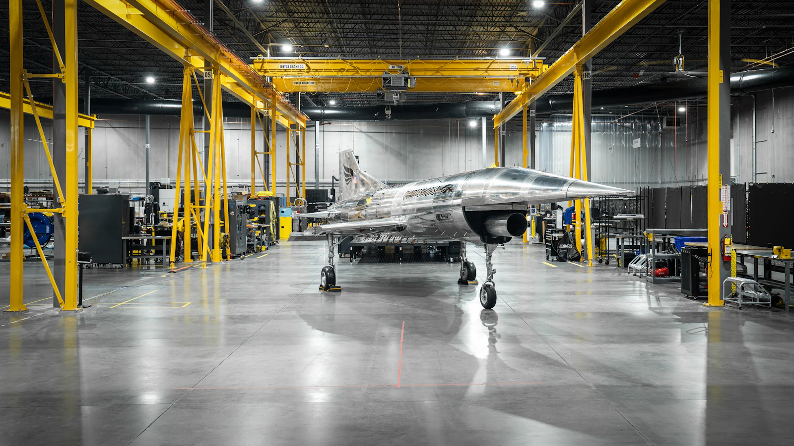 An aircraft on an open shop floor under yellow gantry cranes