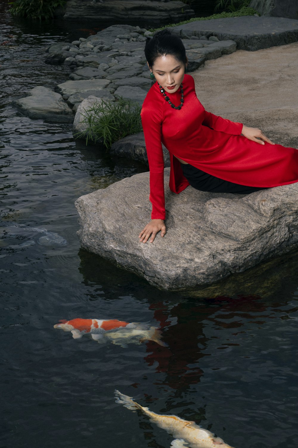 a woman in a red dress sitting on a rock next to a pond