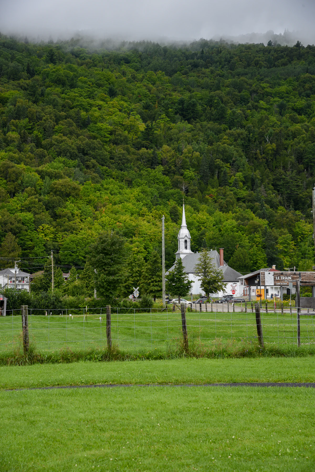 A white church with a steeple at the foot of a forested hillside in Petite-Rivière-Saint-François, with cloud on the ridge