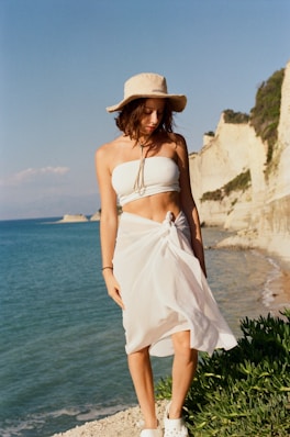 a woman in a white dress and hat standing on a beach