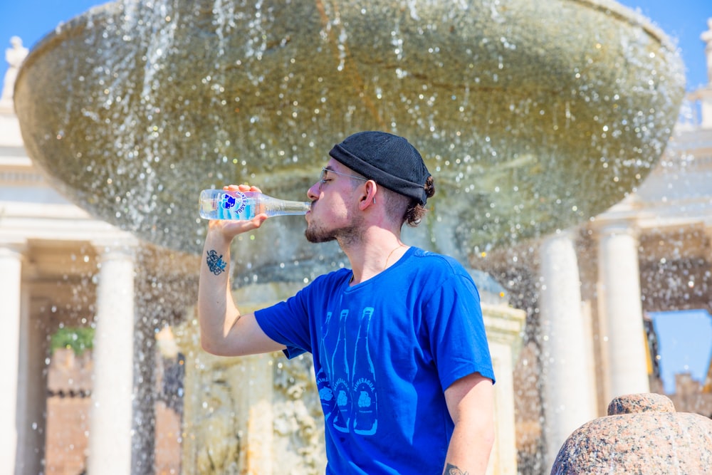 a man drinking water from a bottle in front of a fountain