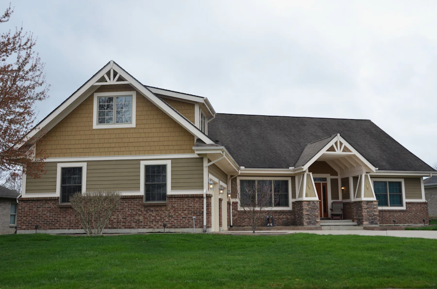 A tan craftsman-style house with a stone base course, gabled entry porch and white trim, standing on a wide sloping front lawn under an overcast sky