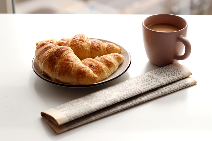 Two butter croissants on a plate beside a cup of coffee and a folded newspaper