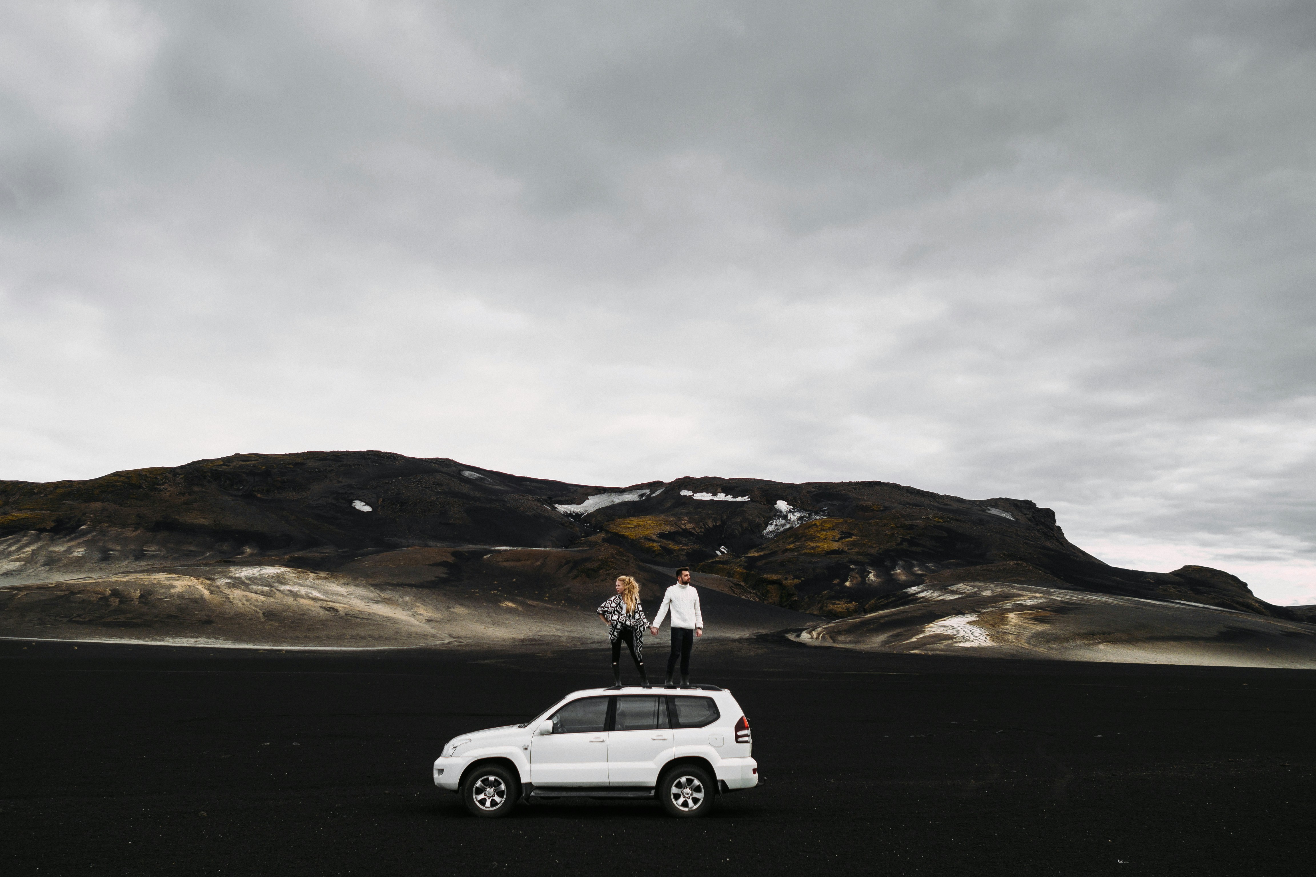two people standing on top of a white car, Lover couple standing by the hand on the roof of the SUV in the Highlands of Iceland.