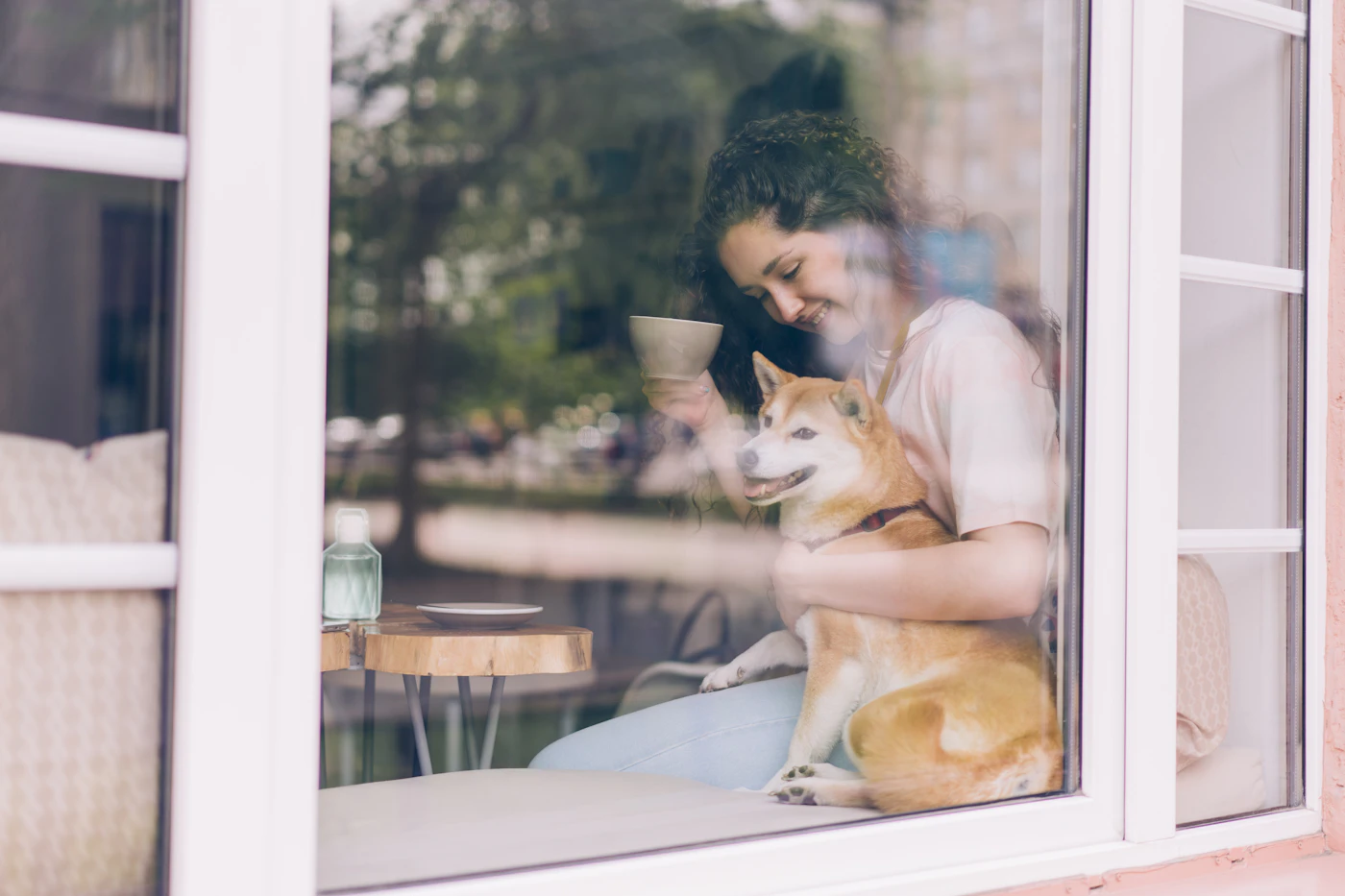 A woman sitting on a sunlit window sill at home holding her dog close, the calm trust of a long partnership