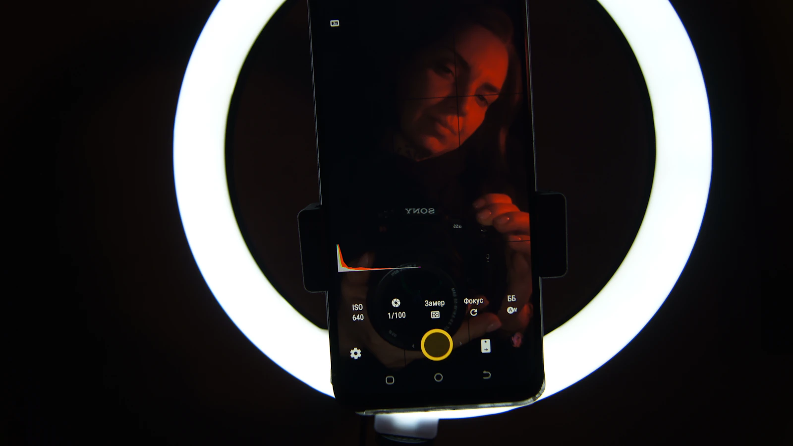 A woman filming herself with a smartphone held in front of a bright ring light in a dark room