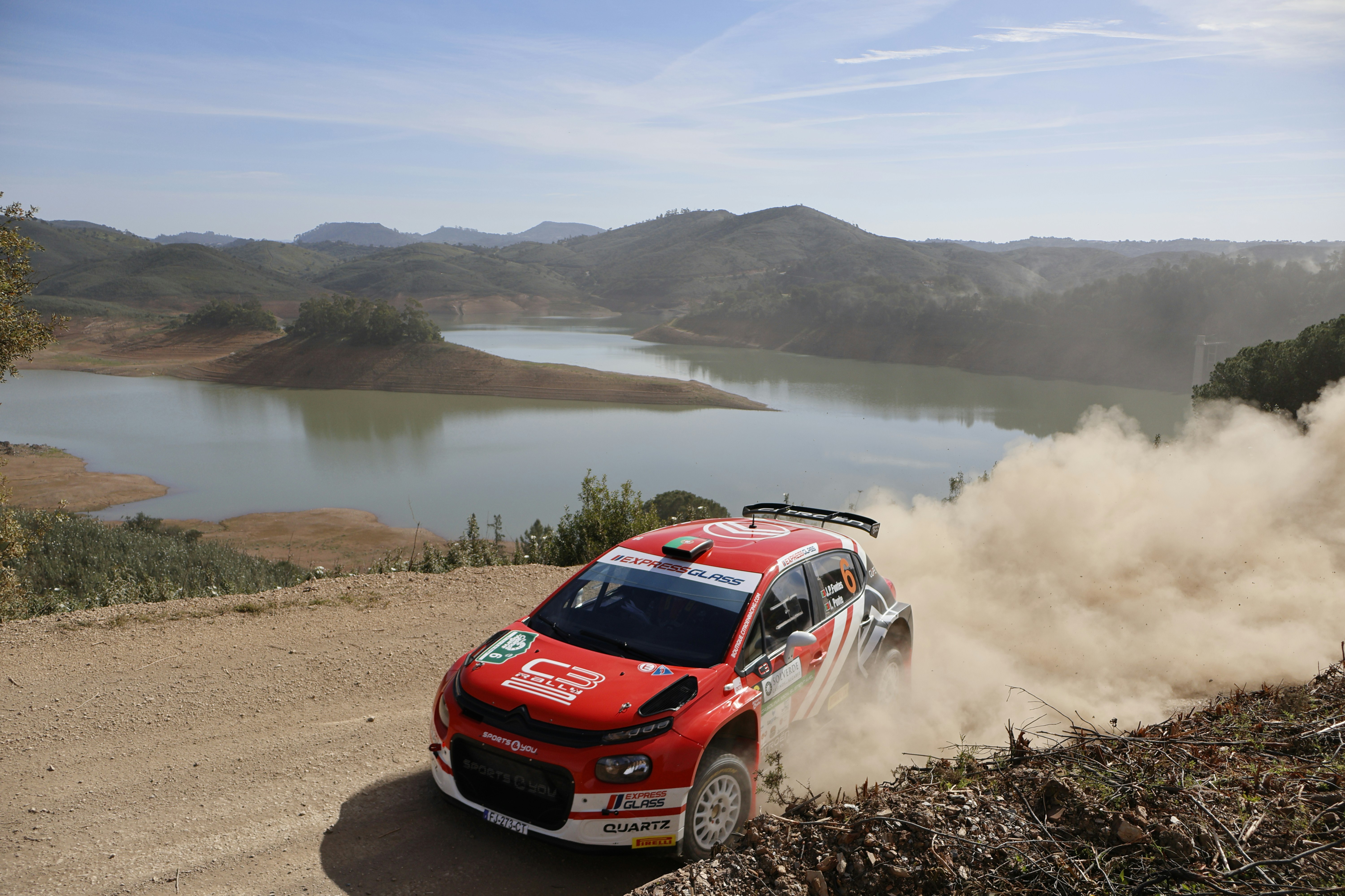 a red car driving down a dirt road next to a lake