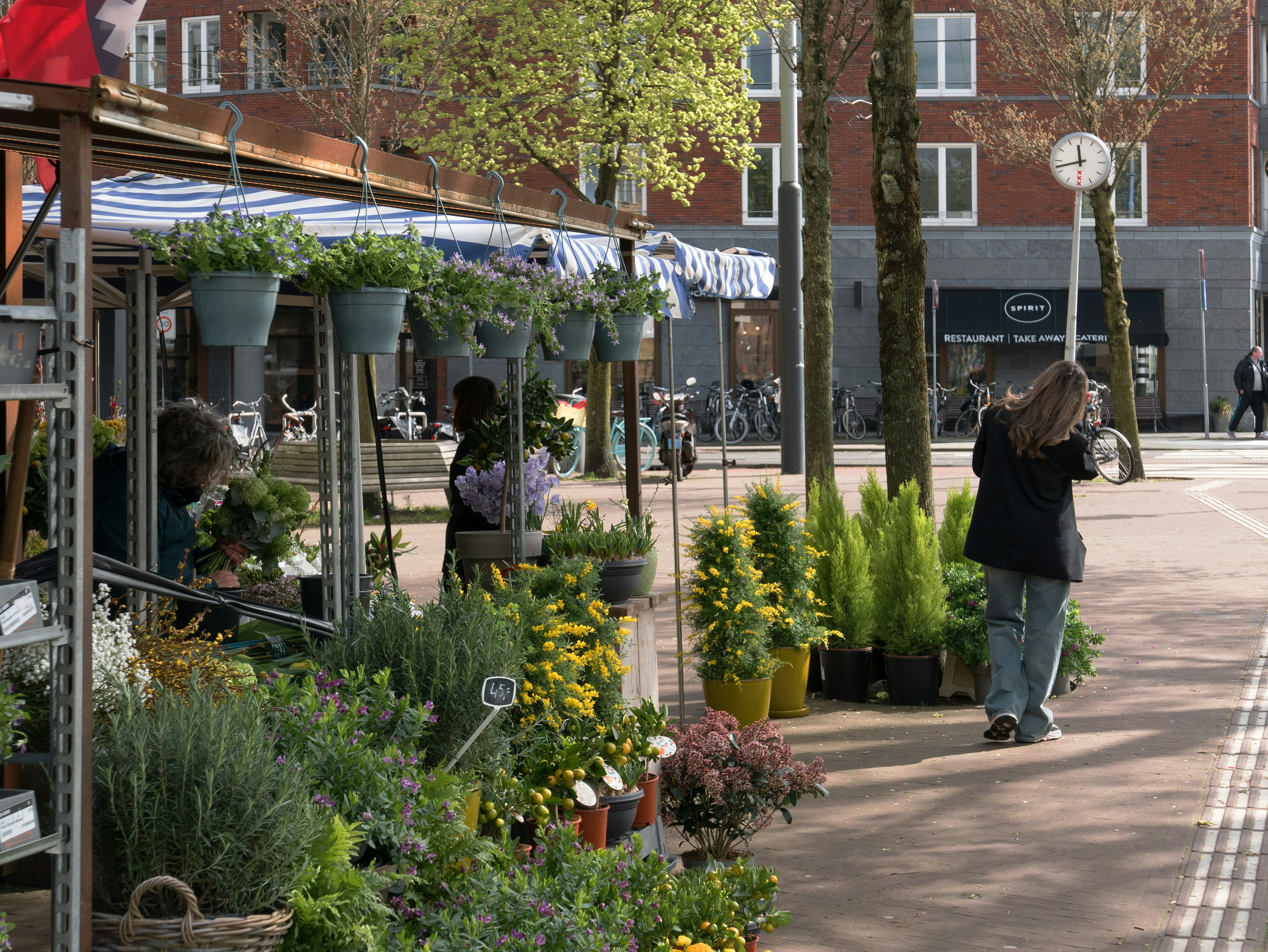 a woman taking a picture of a flower shop, Photo of a flower shop at the corner of the street with a wide sidewalk and people shopping flowers. It is early Spring in town. Street photography of Amsterdam city by Fons Heijnsbroek; free download urban photo, The Netherlands. This street image I share in suitable high resolution for making a nice art print or wallpaper.