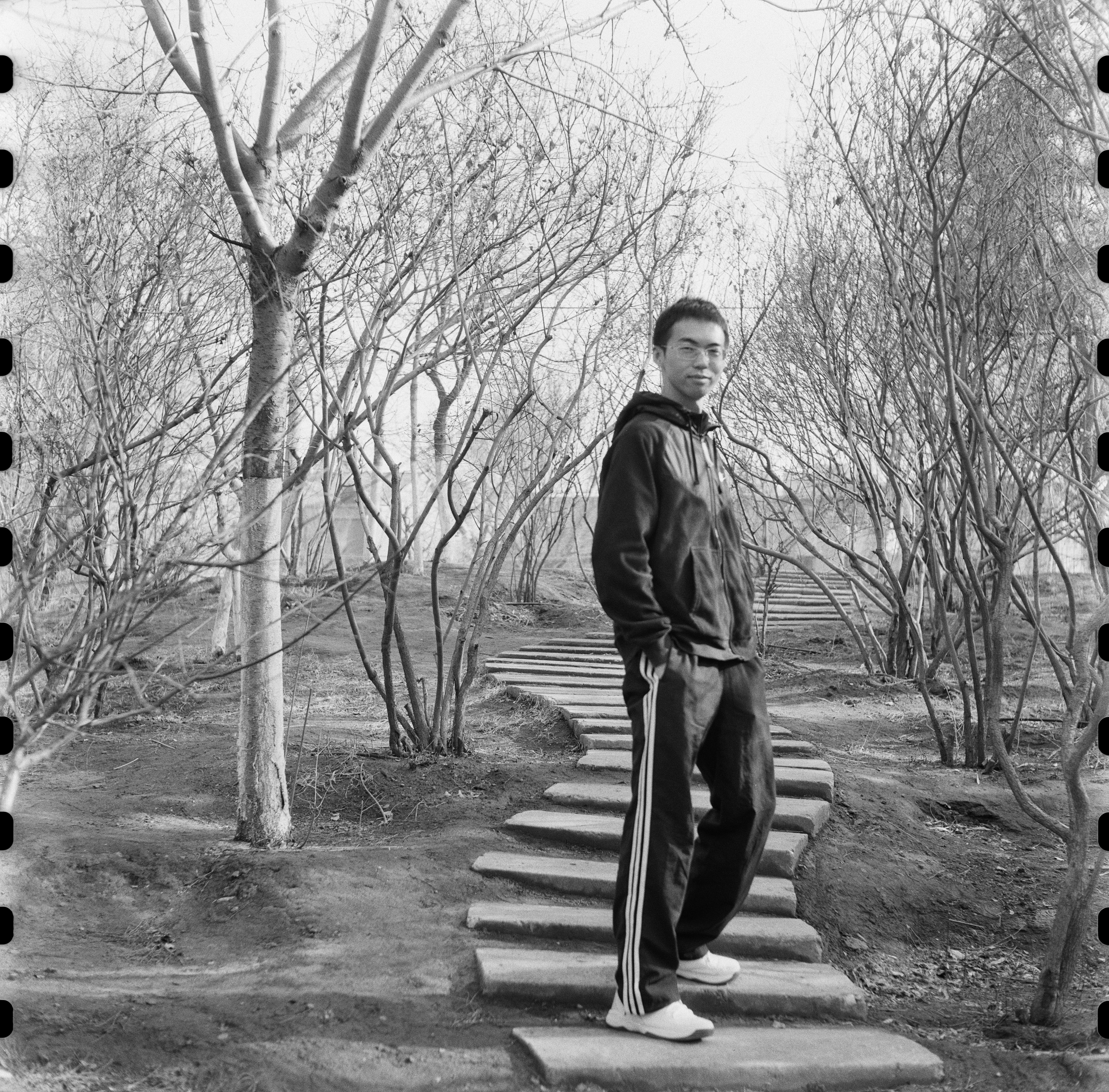 Black-and-white photograph of a man standing on stone steps flanked by bare trees, looking toward the camera.