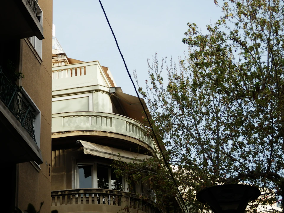 The image depicts a two-story building with a curved balcony on the second floor, situated between two other buildings. The building has white walls and a brown roof, creating an interesting contrast against the clear blue sky. A power line runs across the top of the photo, adding another element to the urban landscape.