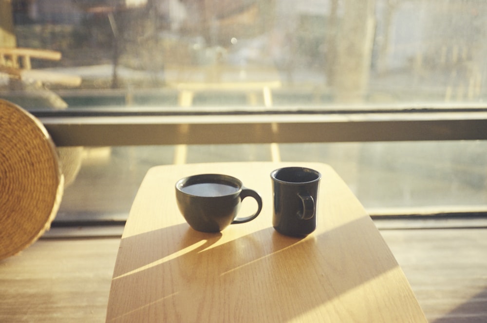 duas xícaras de café sentam-se em uma mesa em frente a uma janela