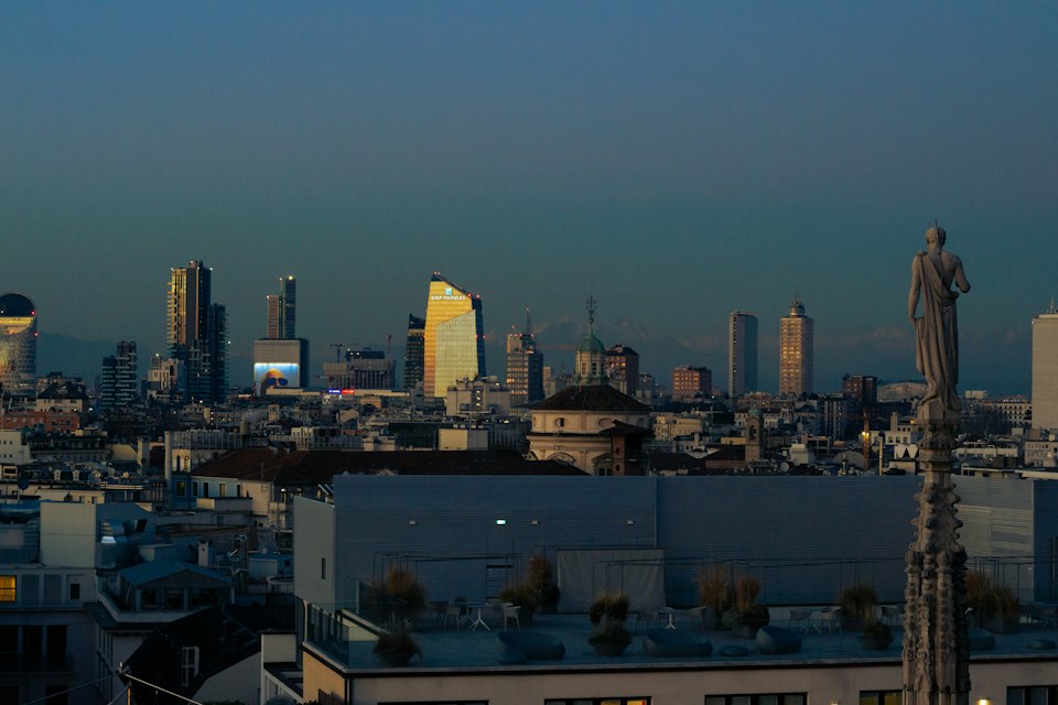 Milan's skyline and a rooftop statue silhouetted at dusk, seen from a terrace