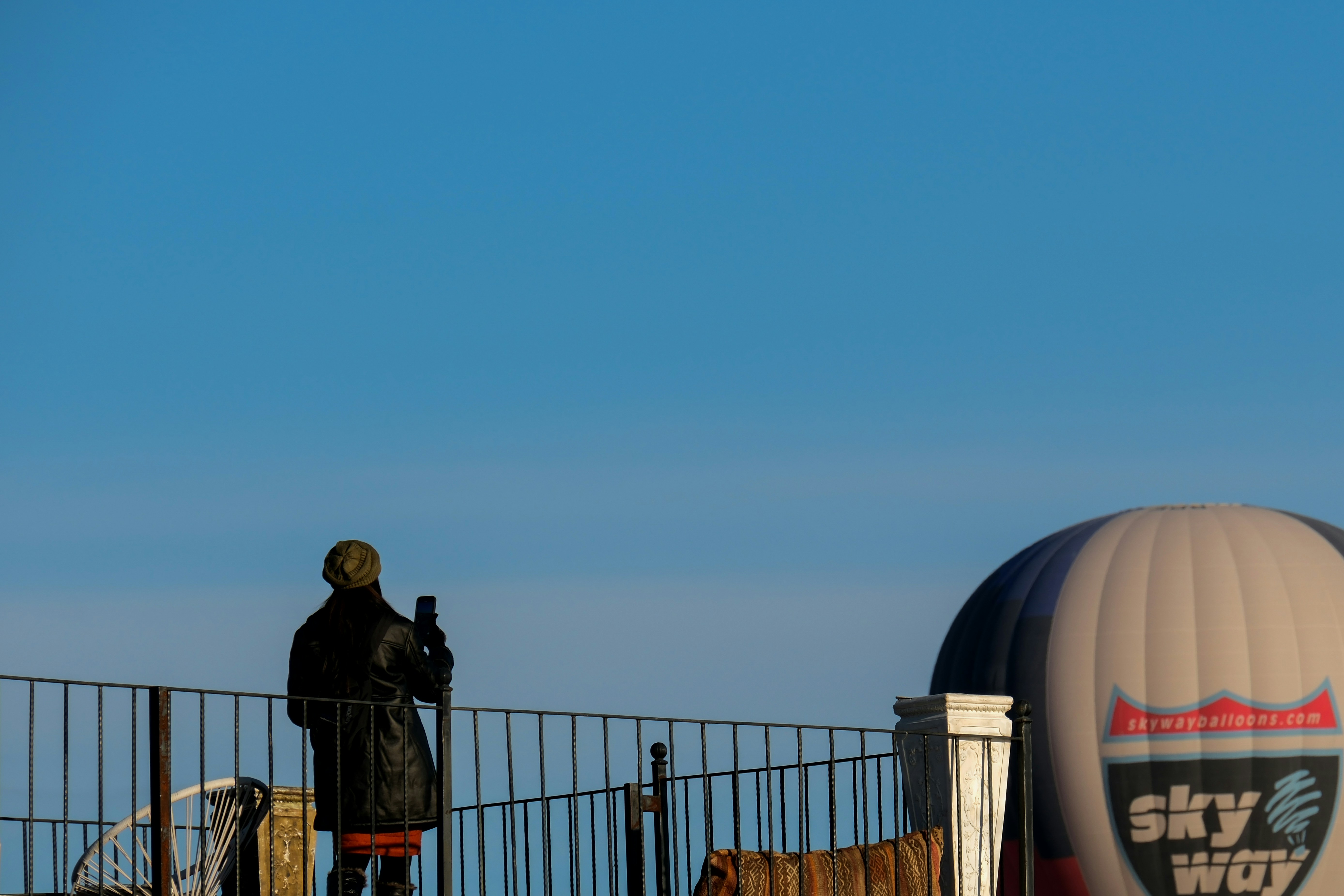a man standing on top of a metal fence next to a hot air balloon - a-man-standing-on-top-of-a-metal-fence-next-to-a-hot-air-balloon-tfZsOSMSVI0