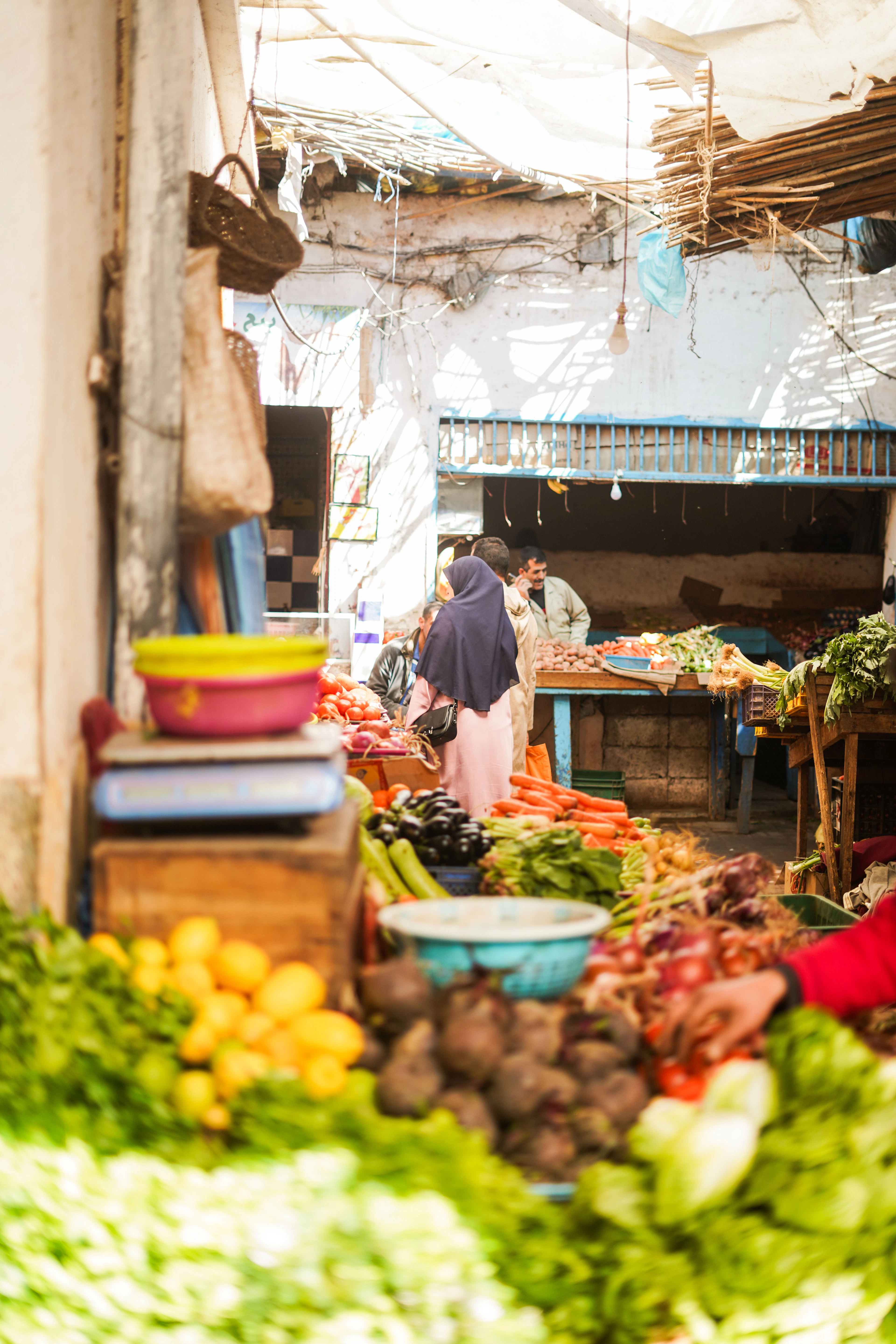 A produce stall in the Essaouira souk heaped with lemons, beetroot, carrots, chard and red onions, a shopper in a plum headscarf standing at the counter under the market's canvas roof