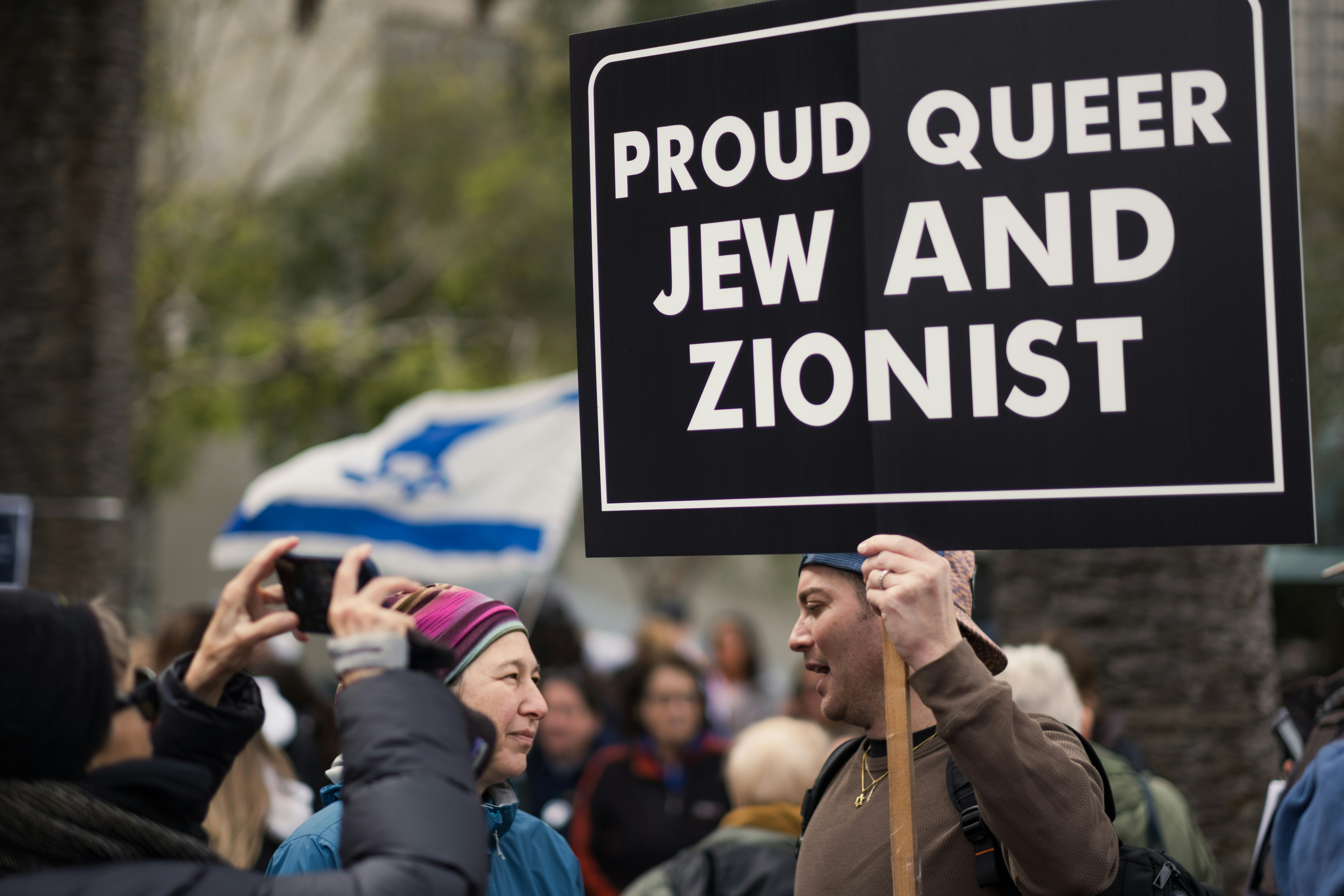 a group of people holding up a sign, At a march against antisemitism, a demonstrator holds up a sign saying, "PROUD QUEER JEW AND ZIONIST"