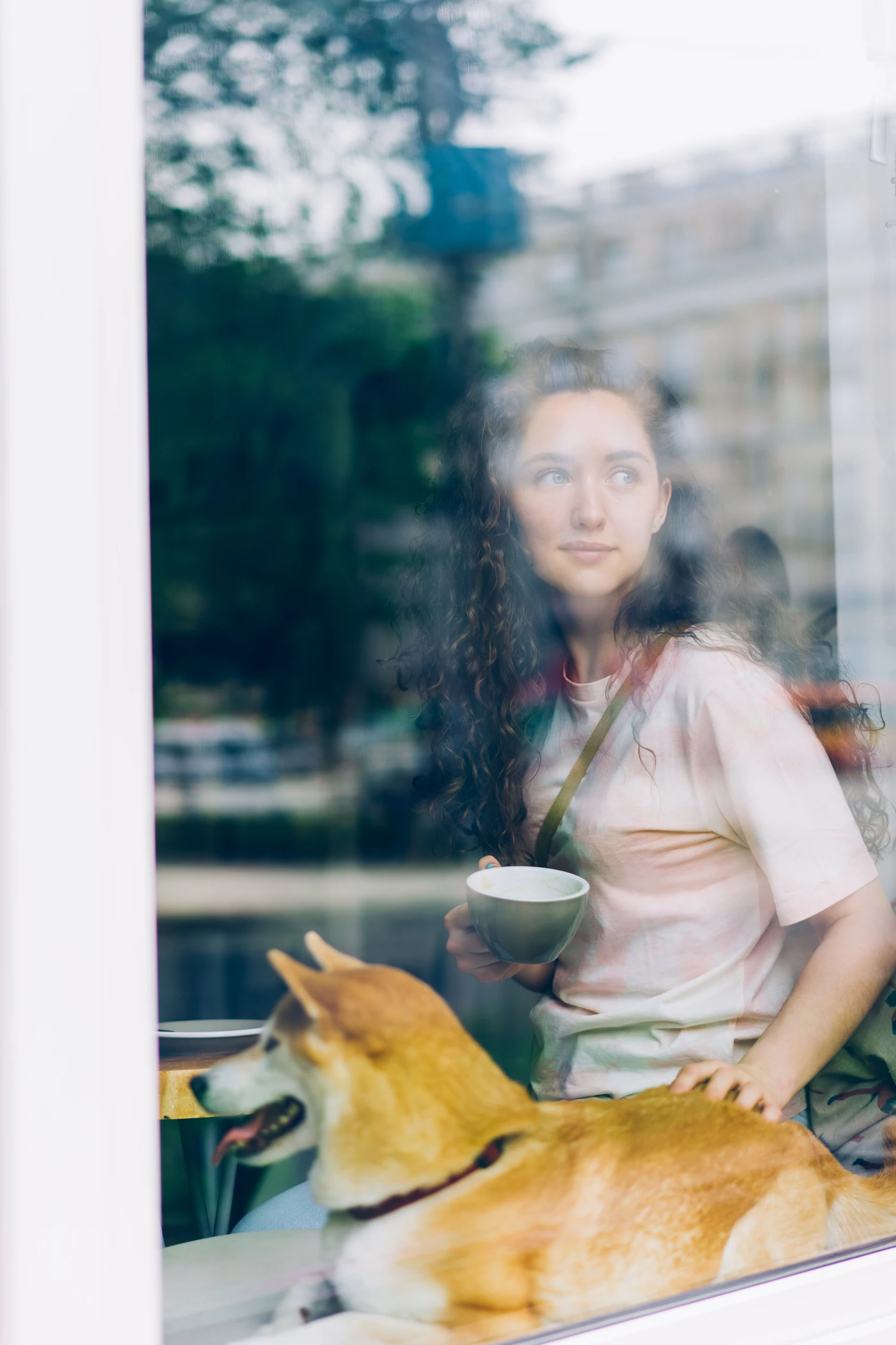 A woman with a warm drink resting a hand on a calm dog by a sunlit window