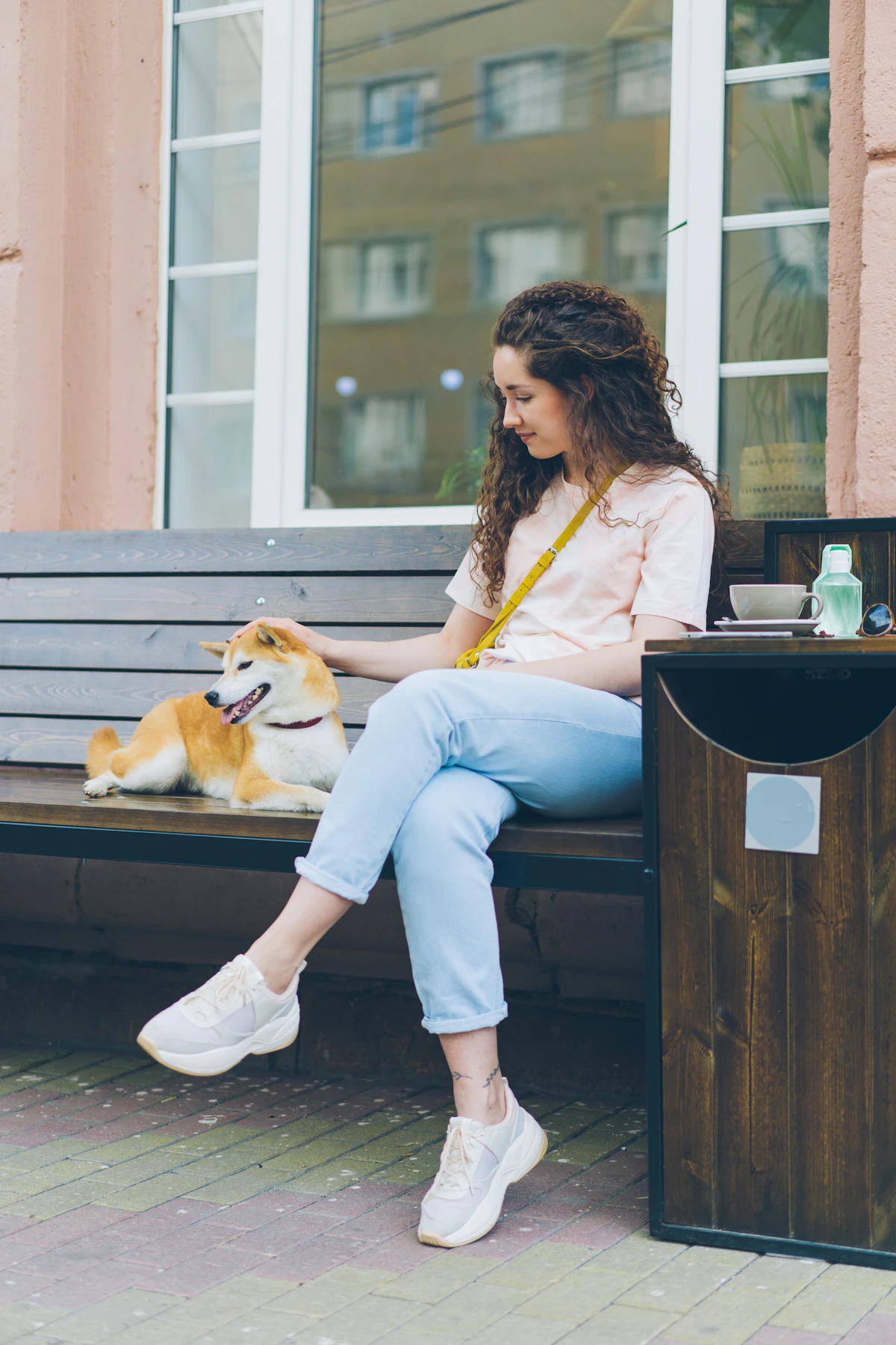 A young woman in jeans sits on an outdoor bench gently petting a Shiba Inu dog resting beside her outside a building